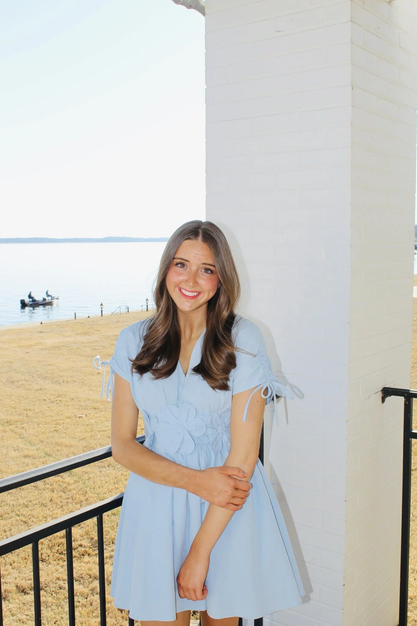 A young woman with long brown hair and a bright smile, wearing a light blue dress with floral embroidery, standing outdoors next to a white brick wall, with a lake, boat, and grassy area in the background.