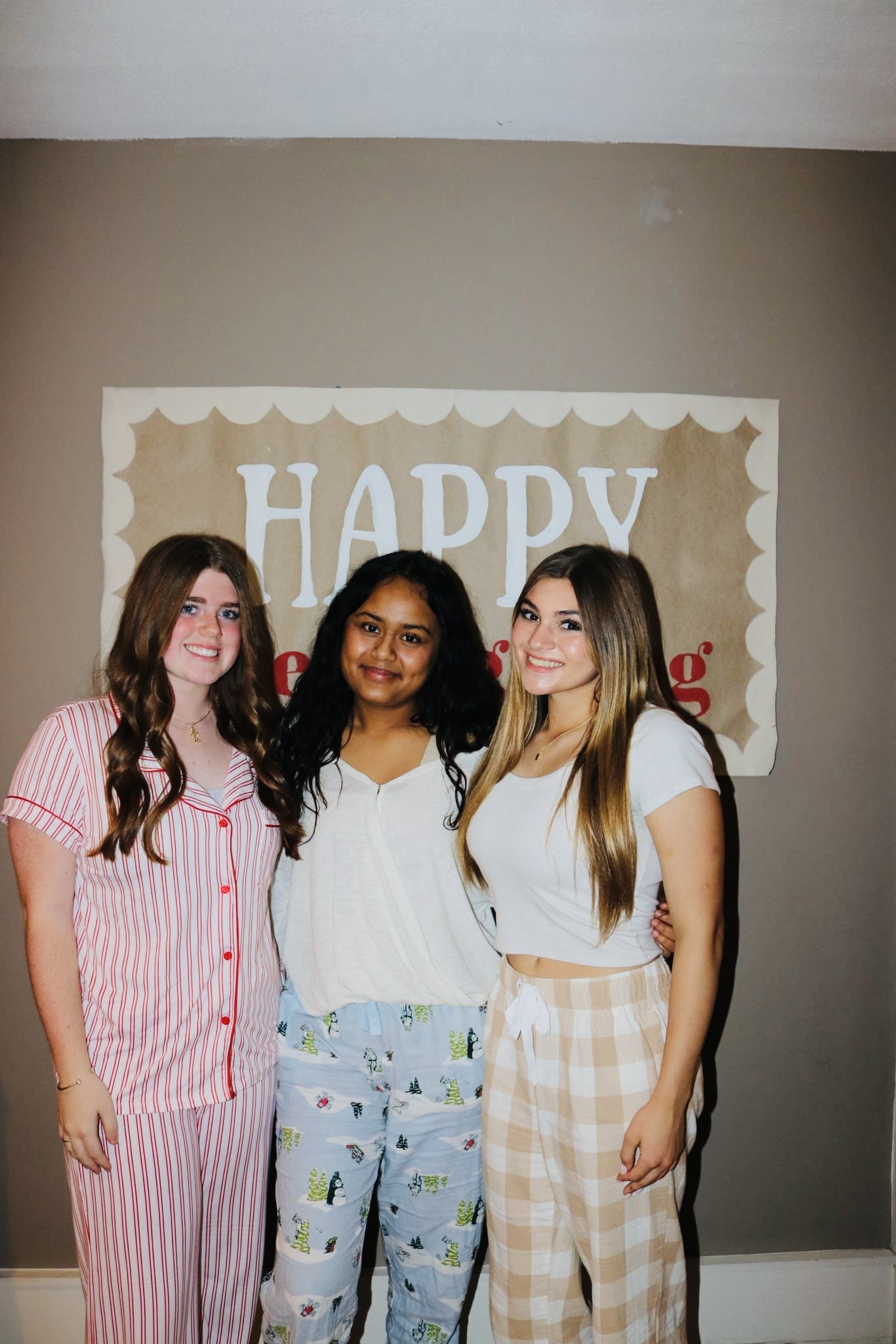 Three women standing together in front of a beige wall with a 'HAPPY' banner, smiling at the camera in pajamas.