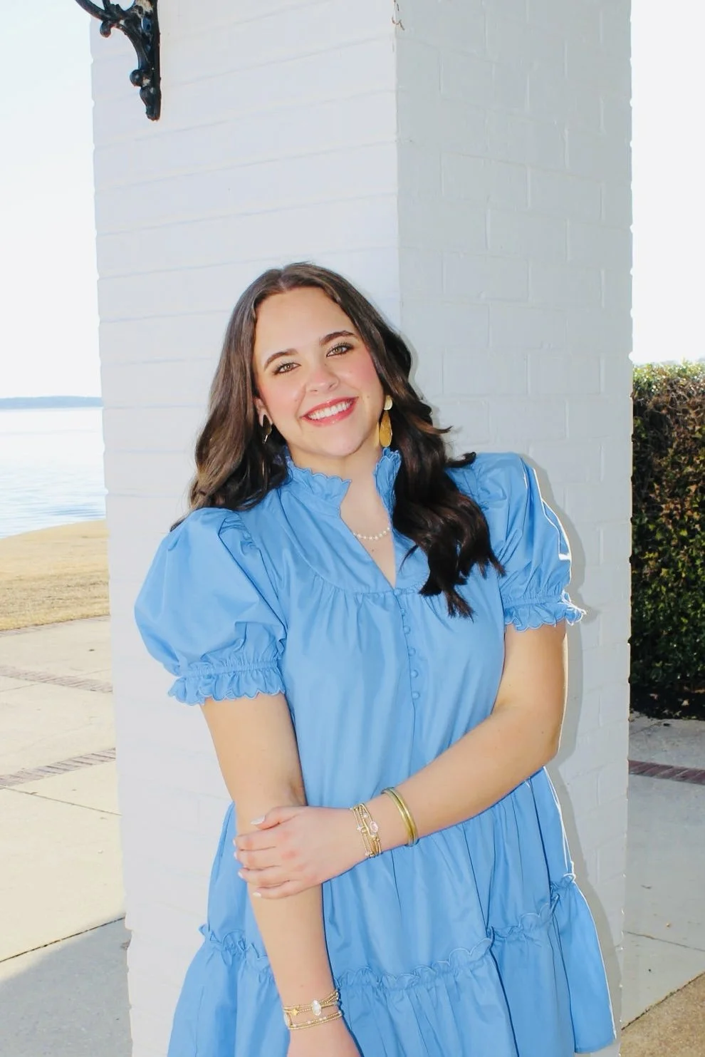 Young woman smiling outdoors, wearing a bright blue dress with puffed sleeves, standing near a white brick wall with a body of water visible in the background.