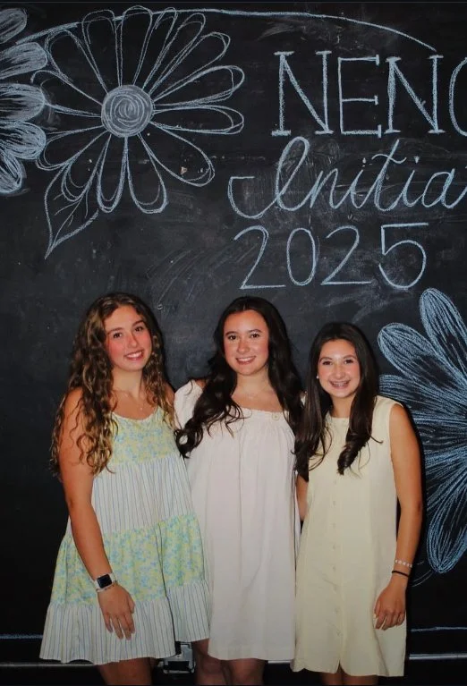 Three young women standing in front of a black chalkboard with drawings of flowers and butterflies, and text that reads 'NENA graduation 2025'.