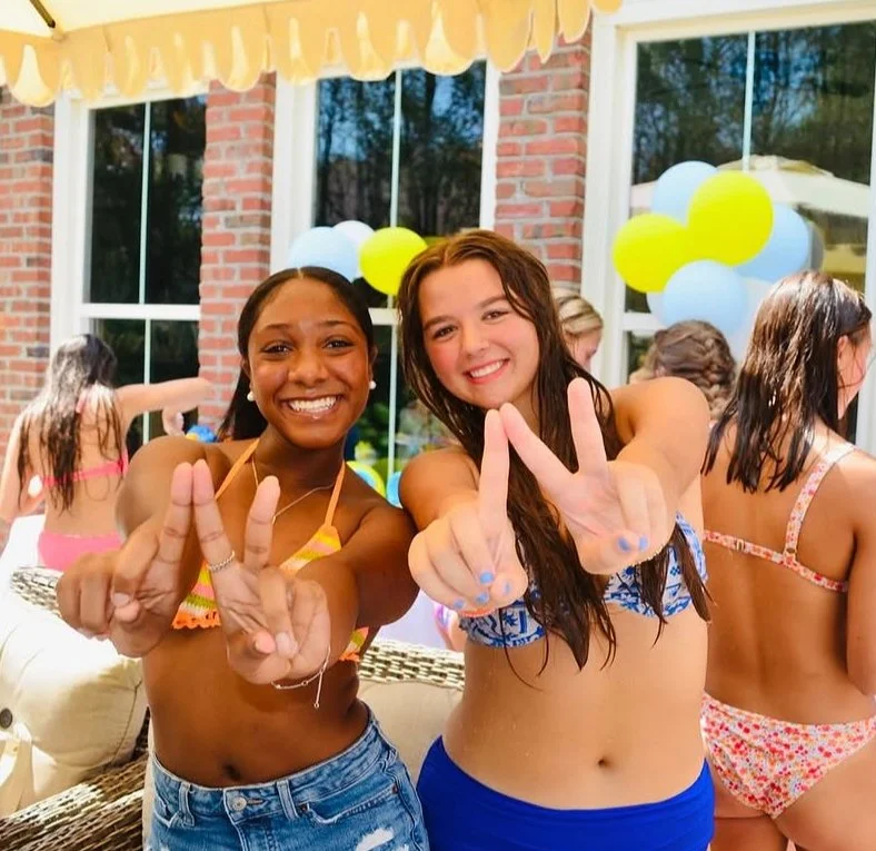Two girls in swimsuits smiling and showing peace signs at a pool party, with balloons and other people in swimsuits in the background.
