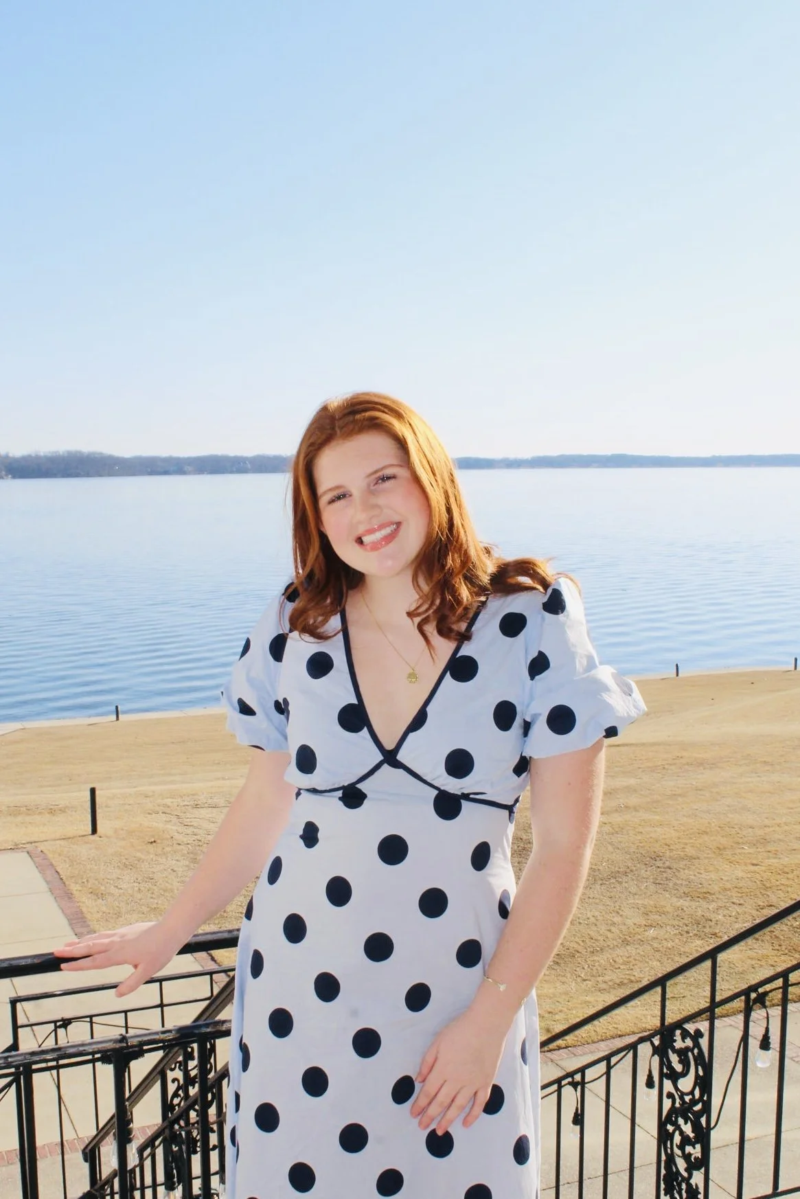 A woman with red hair wearing a white dress with black polka dots, standing on a balcony by a lake with a clear blue sky.