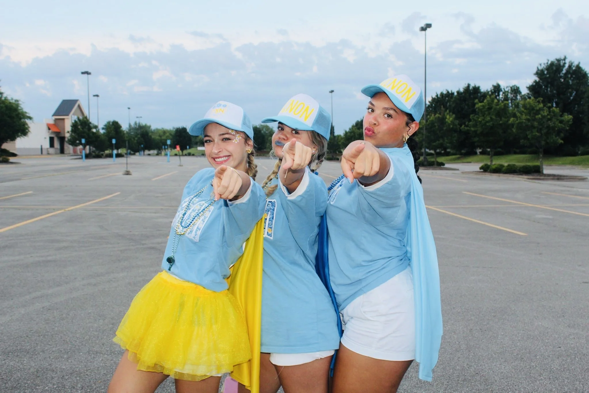 Three young women dressed in blue shirts, yellow skirt, and white shorts with blue cap and superhero capes posing in an empty parking lot.