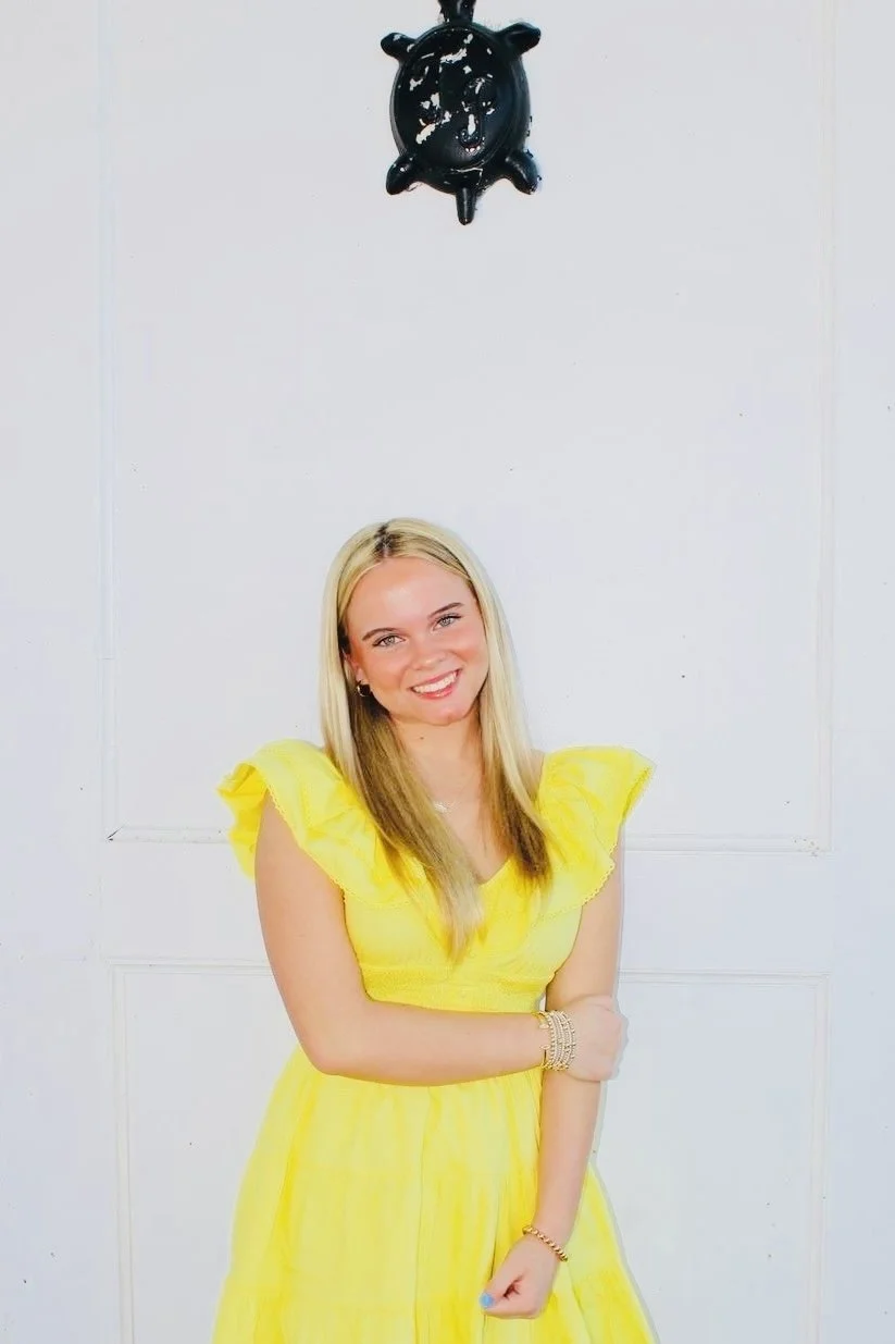 A smiling young woman with blonde hair wearing a yellow dress, standing in front of a white wall with a black decorative piece hanging above her.