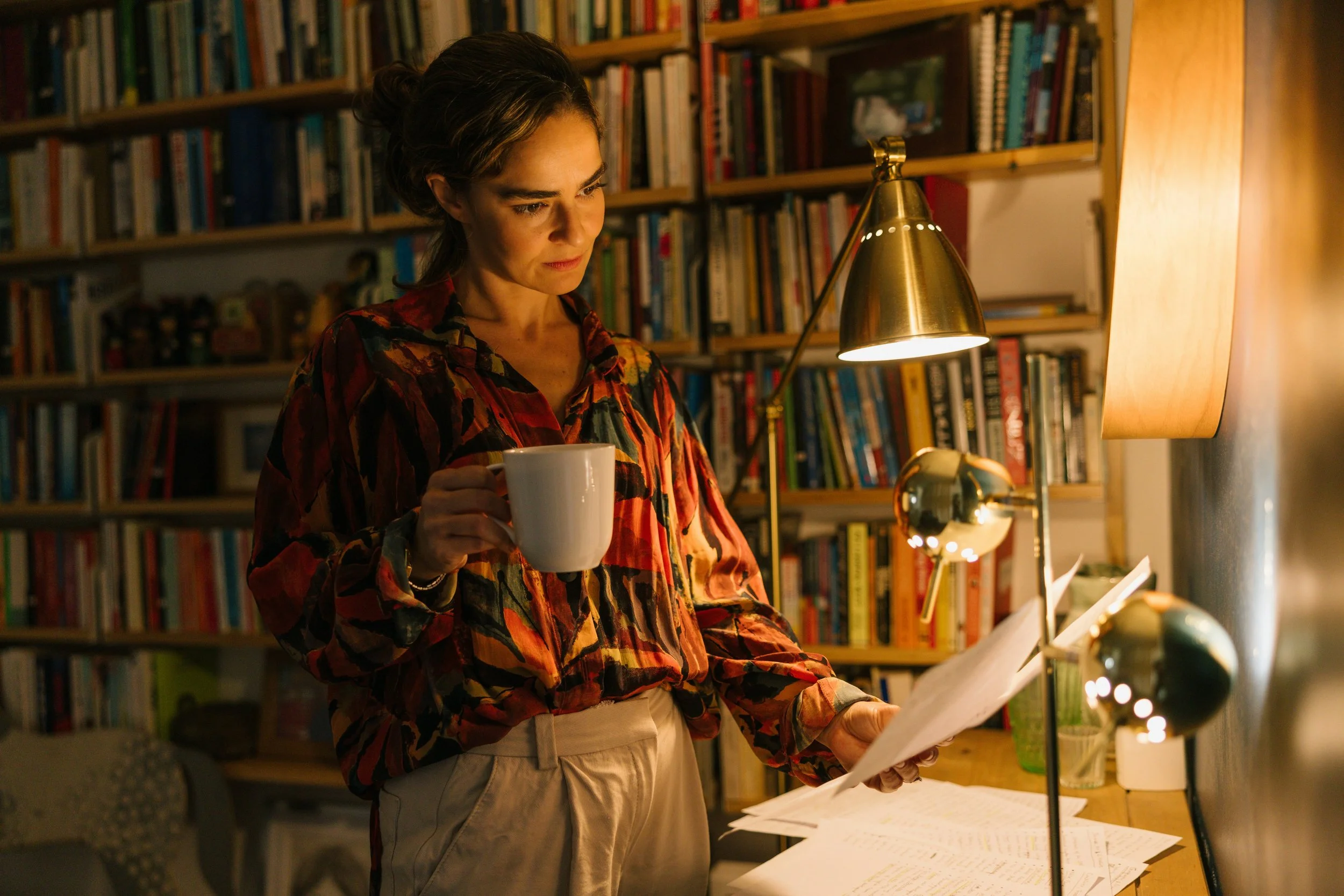 A woman in a patterned blouse holds a cup of coffee in one hand, and a few pages on the other. She is surrounded by books, and reading under a lamp light.