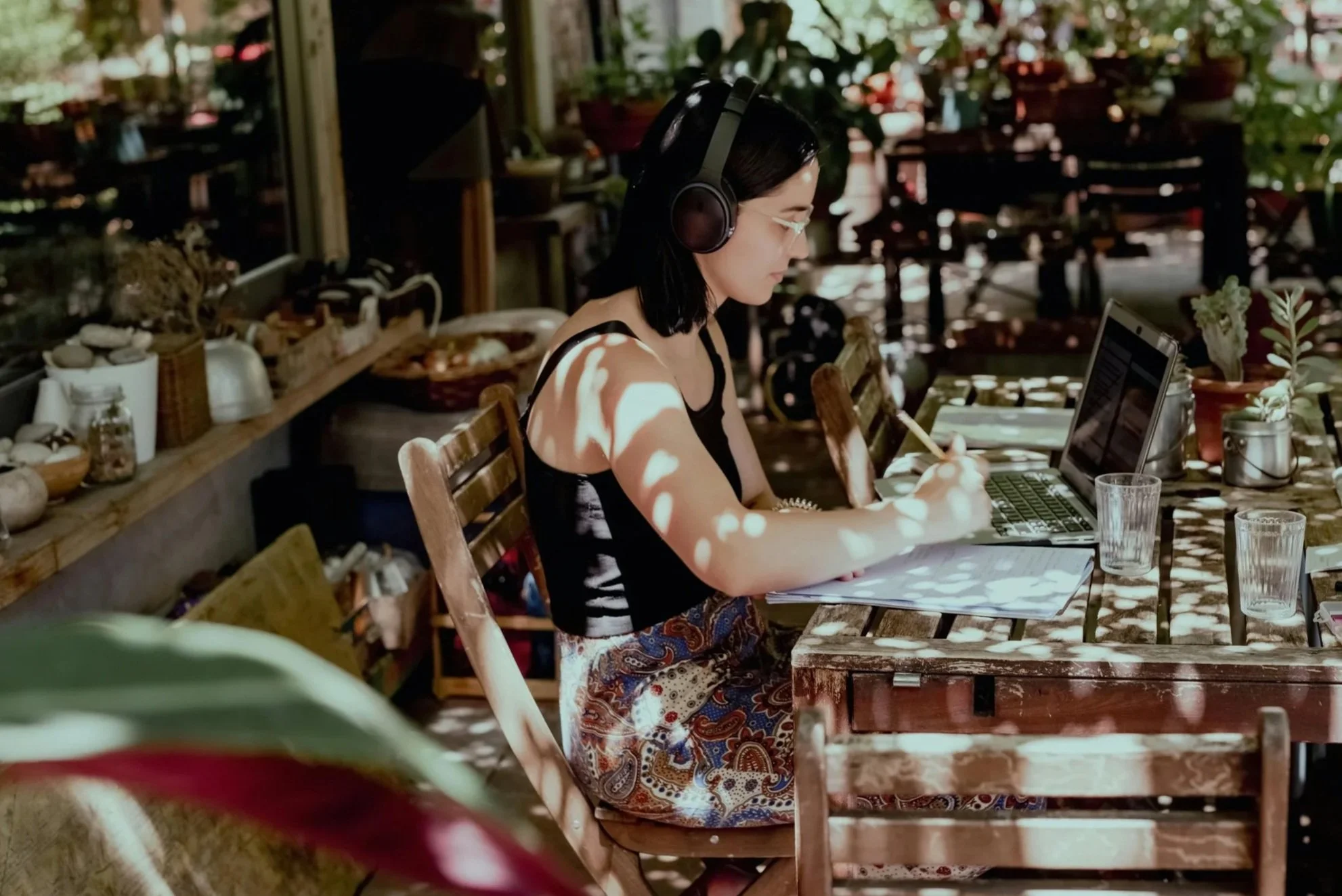 A woman wearing headphones sits at a wooden table inside a room filled with plants, on top of which is a laptop and a notebook.