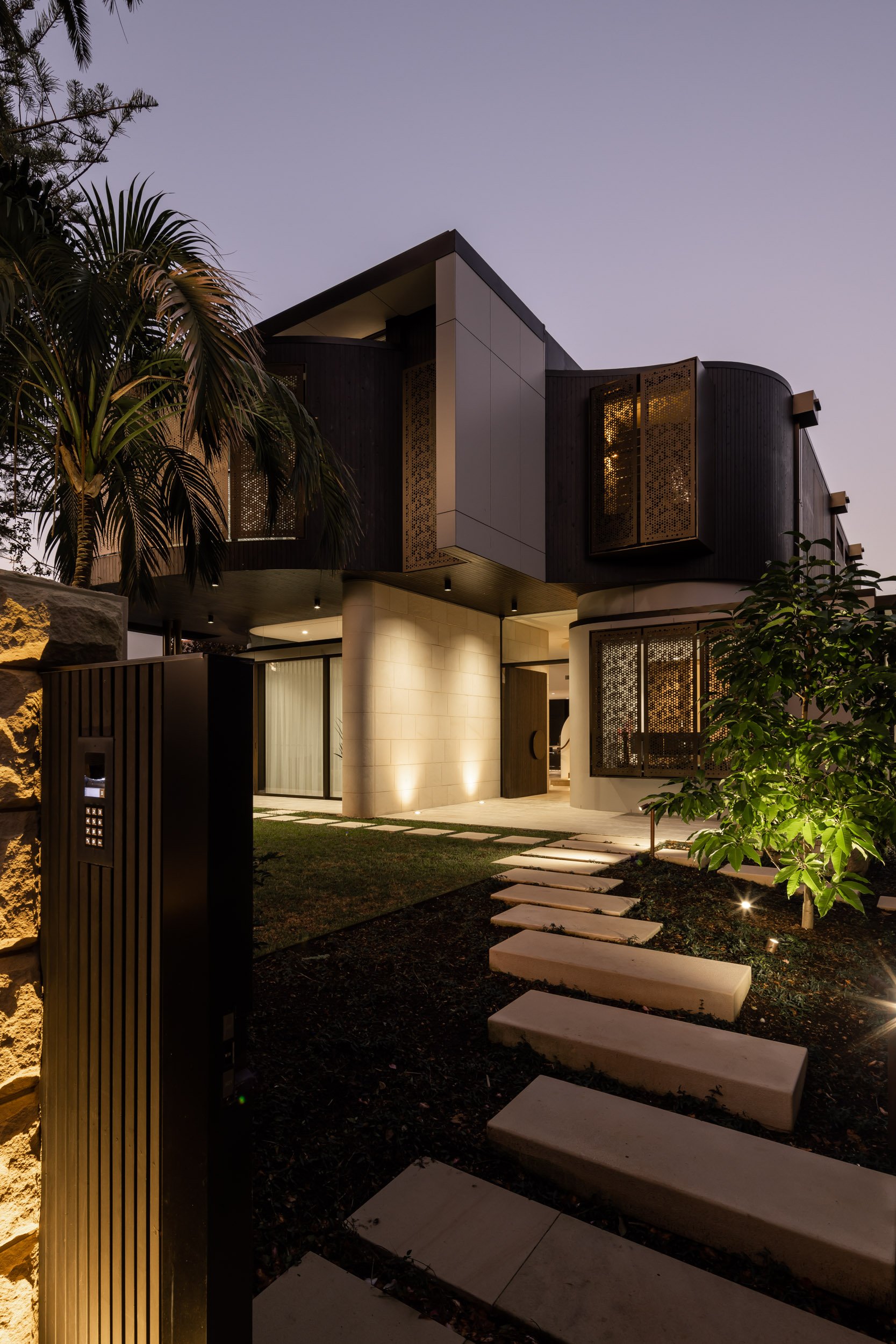 Modern two-story house with illuminated exterior and pathway, surrounded by palm trees at dusk.