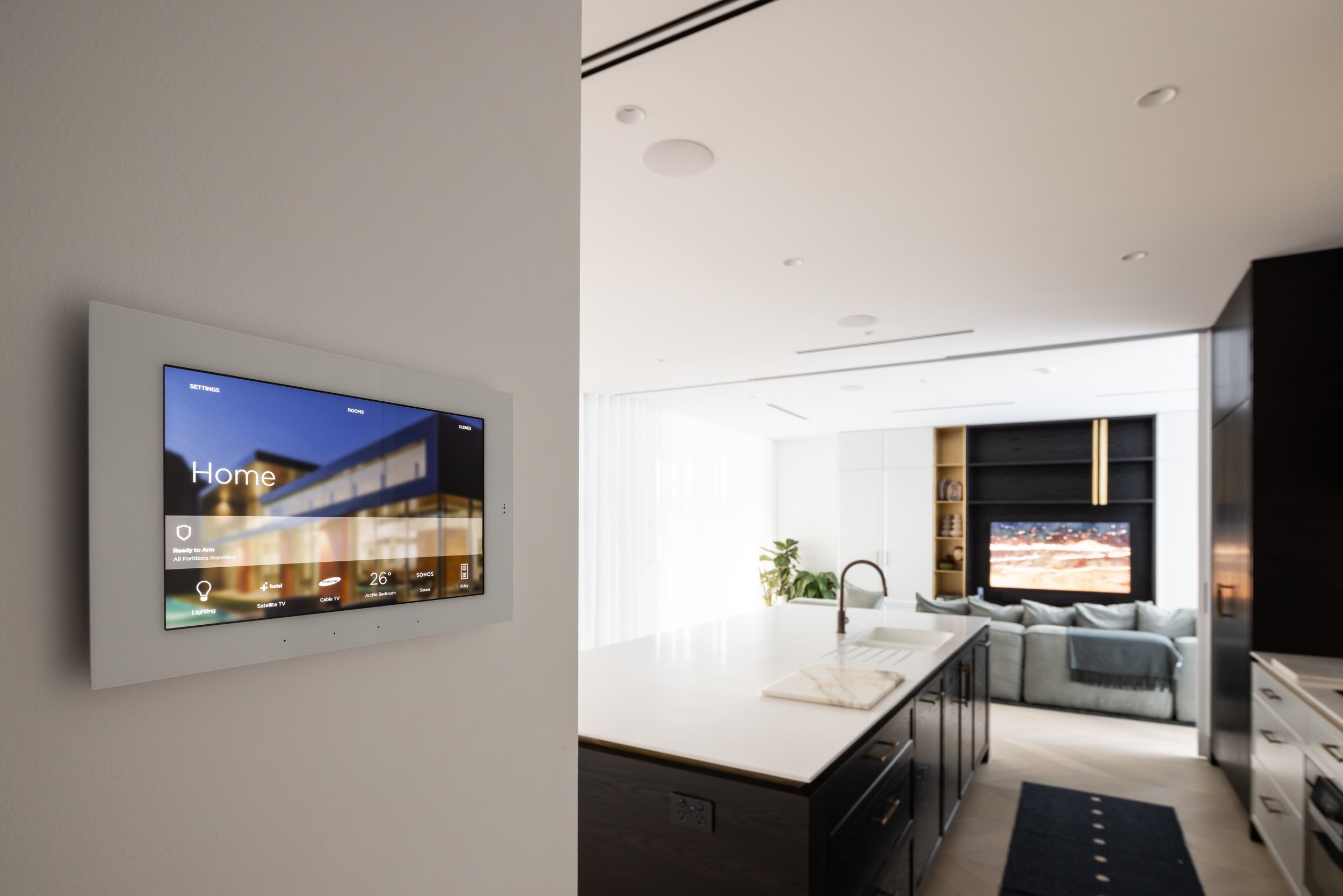 Smart home control panel on wall in modern kitchen leading to living room with TV and sofa.