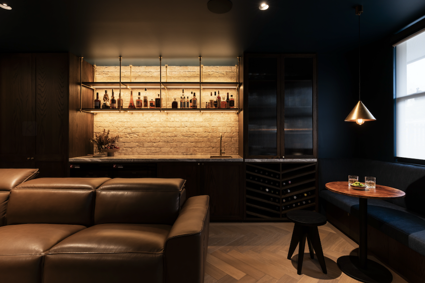 Stylish home bar with leather sofa, backlit shelves displaying liquor bottles, modern pendant light, small round table with drinks, dark wood cabinetry, and exposed brick wall.