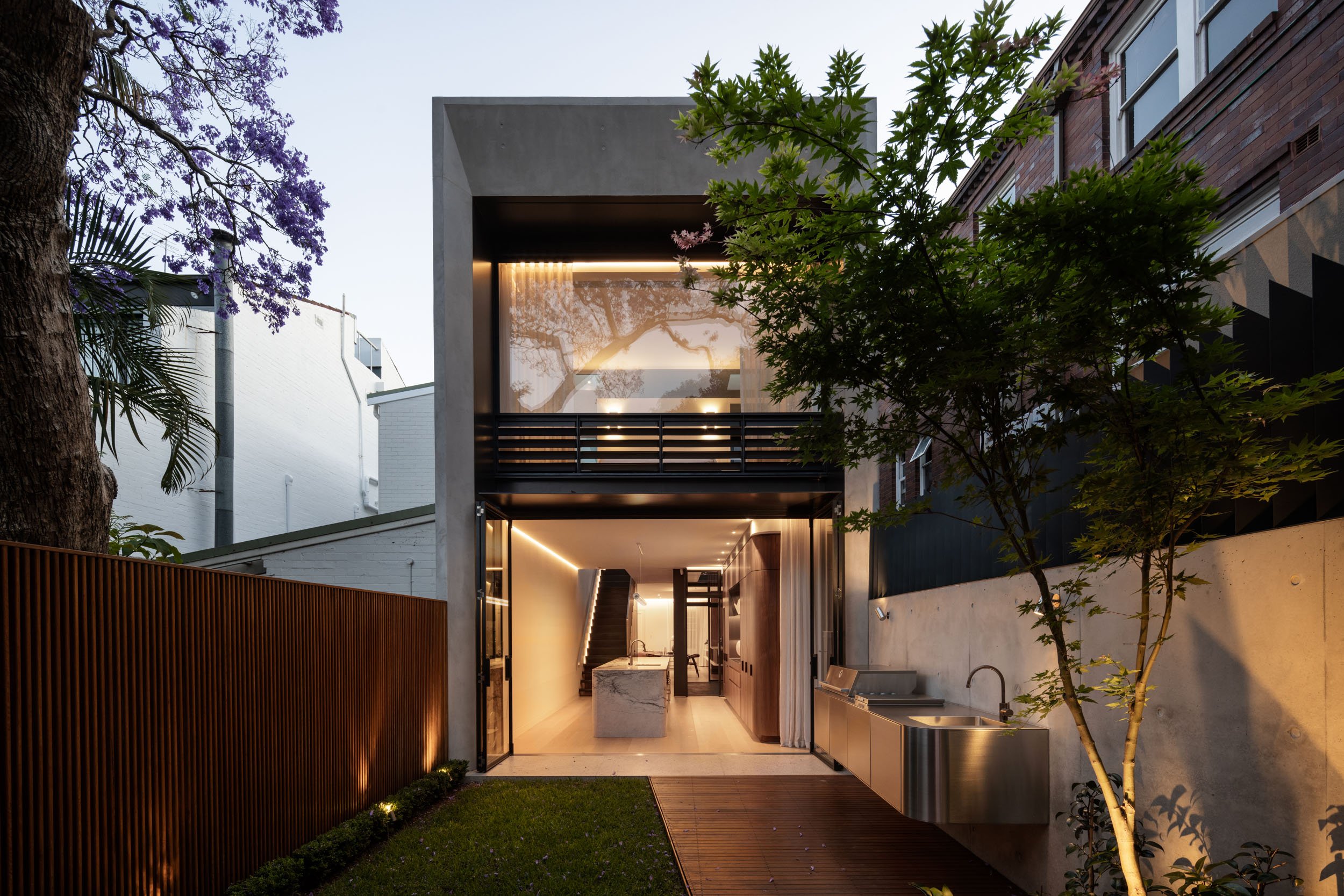 Modern two-story house with large glass windows and open interior, featuring a sleek kitchen with a marble island and an outdoor deck with a built-in barbecue and sink. Surrounded by trees and a wooden fence.