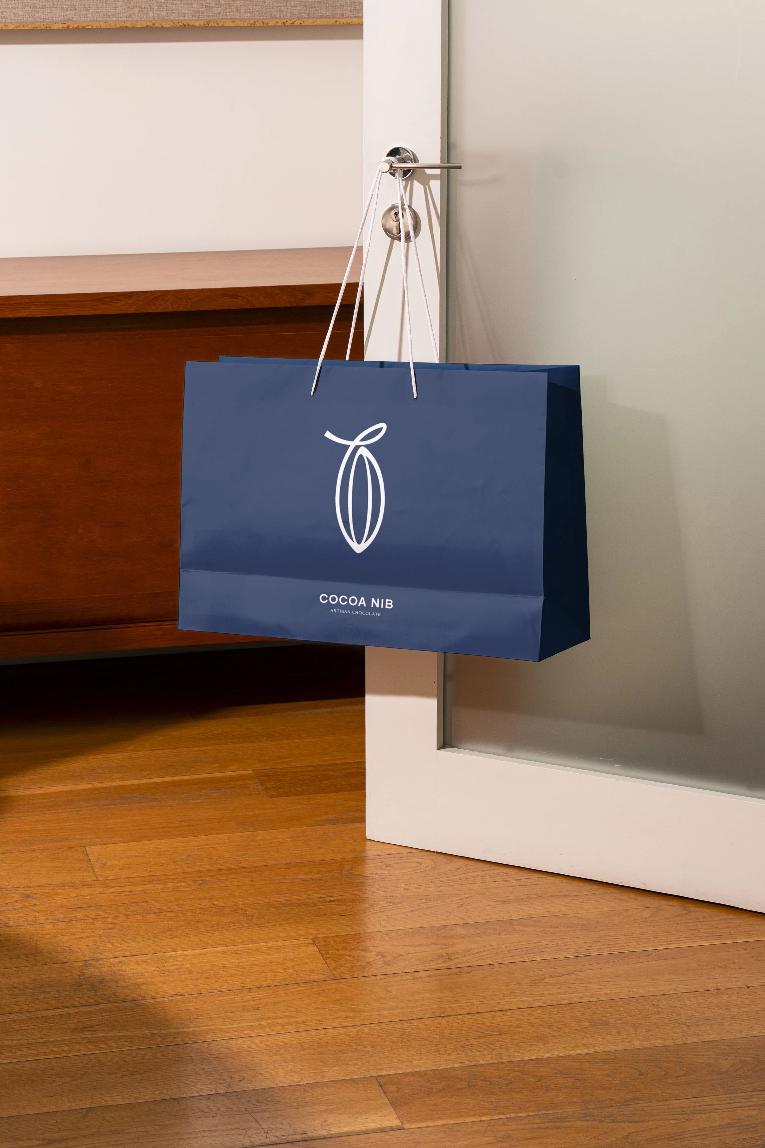 A navy blue shopping bag hanging on a hook next to a white door. The bag has a simple white outline of a cocoa pod and the words "COCOA NIB ARTISAN CHOCOLATE" printed on it.