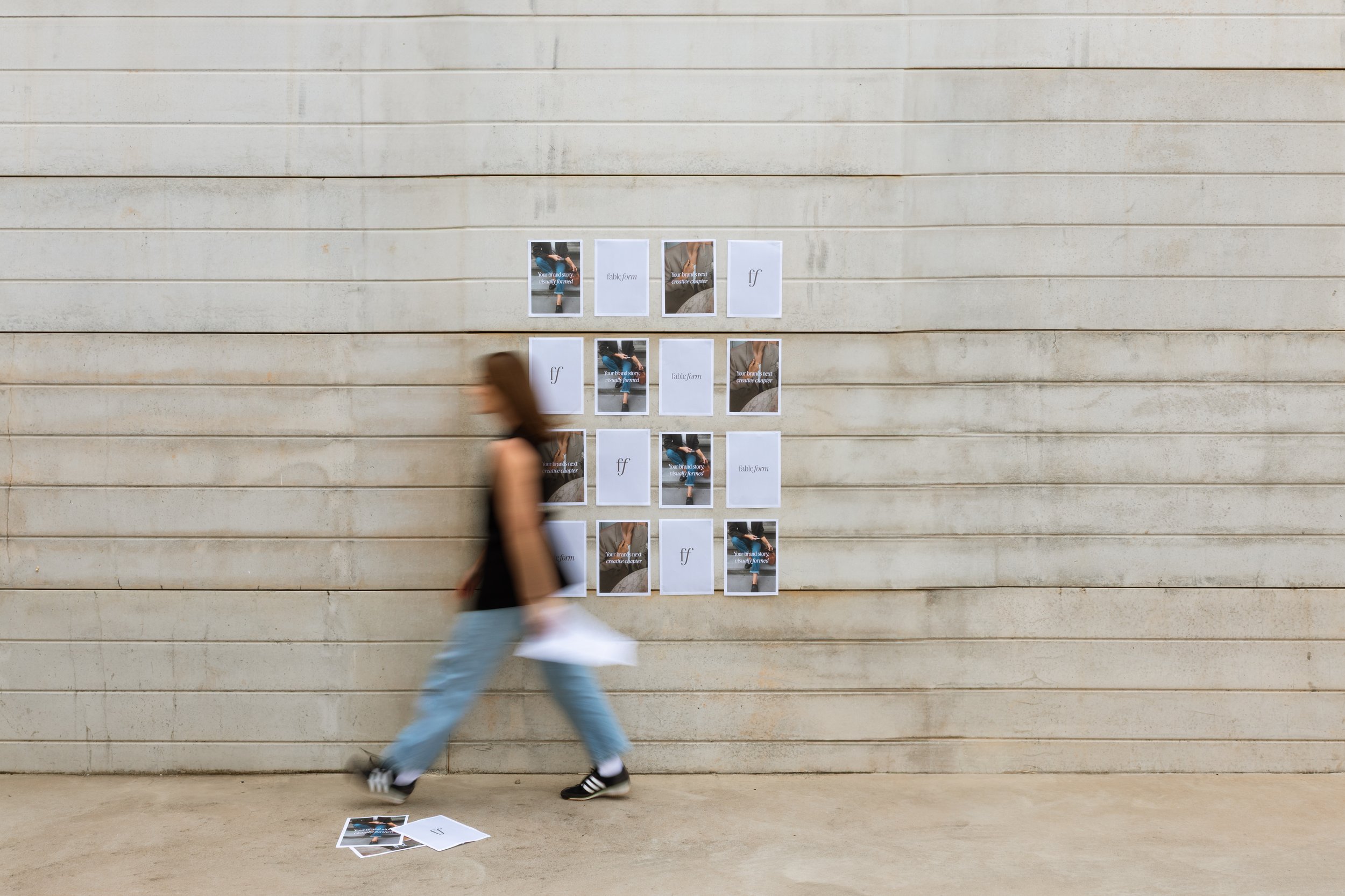 A woman walking past a wall with arranged posters and papers, some on the ground, featuring images and text.