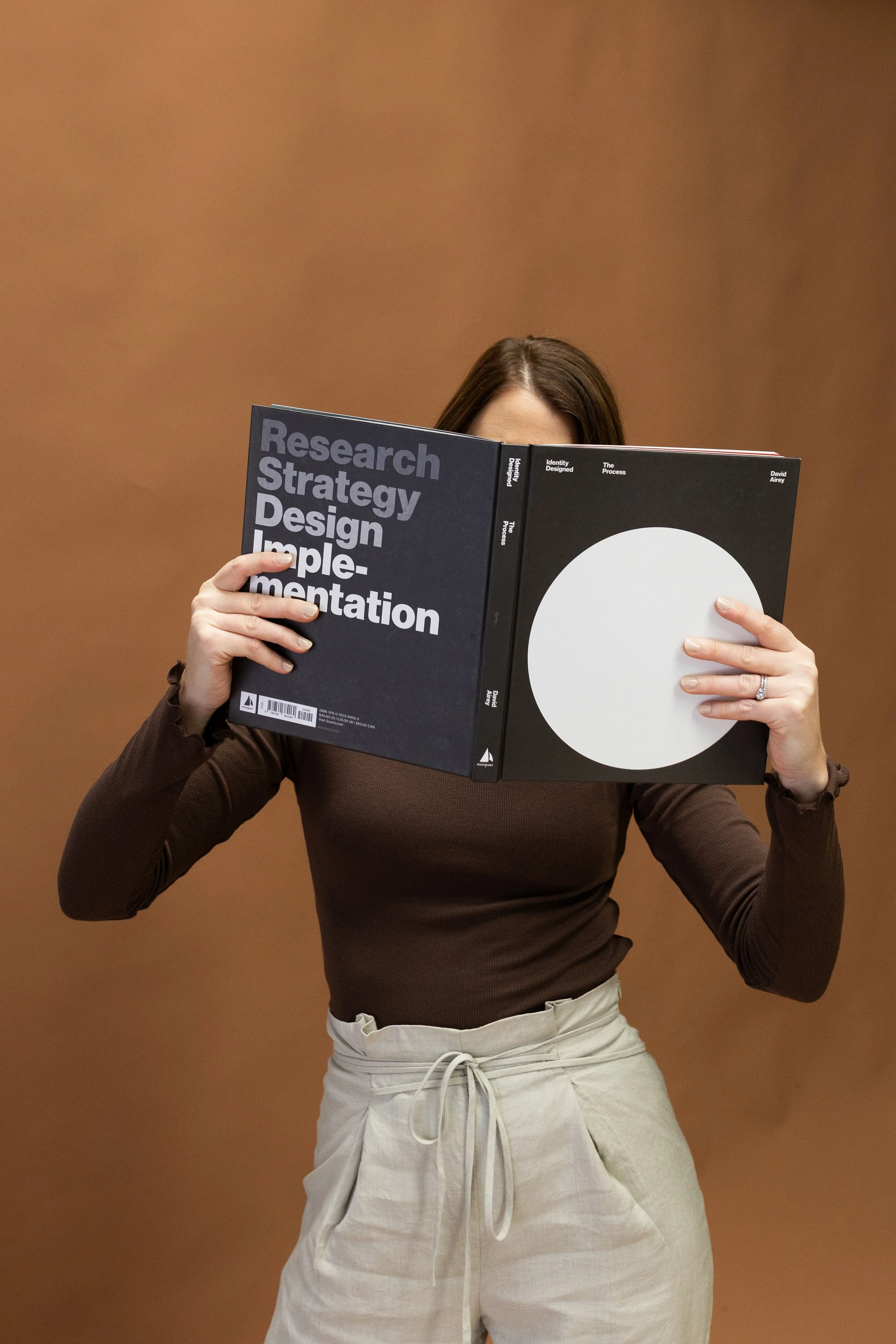 Woman holding an open book titled 'Identity Designed - The Process', with her face obscured behind the pages, standing against a plain brown background.