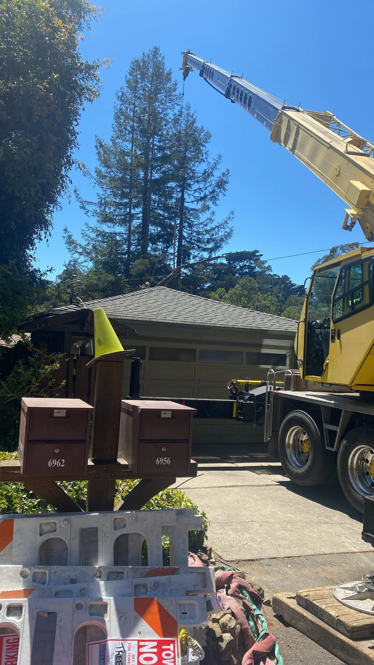 A construction crane lifting a branch or a large object above a residential house with trees. There are utility boxes and construction signs in the foreground.
