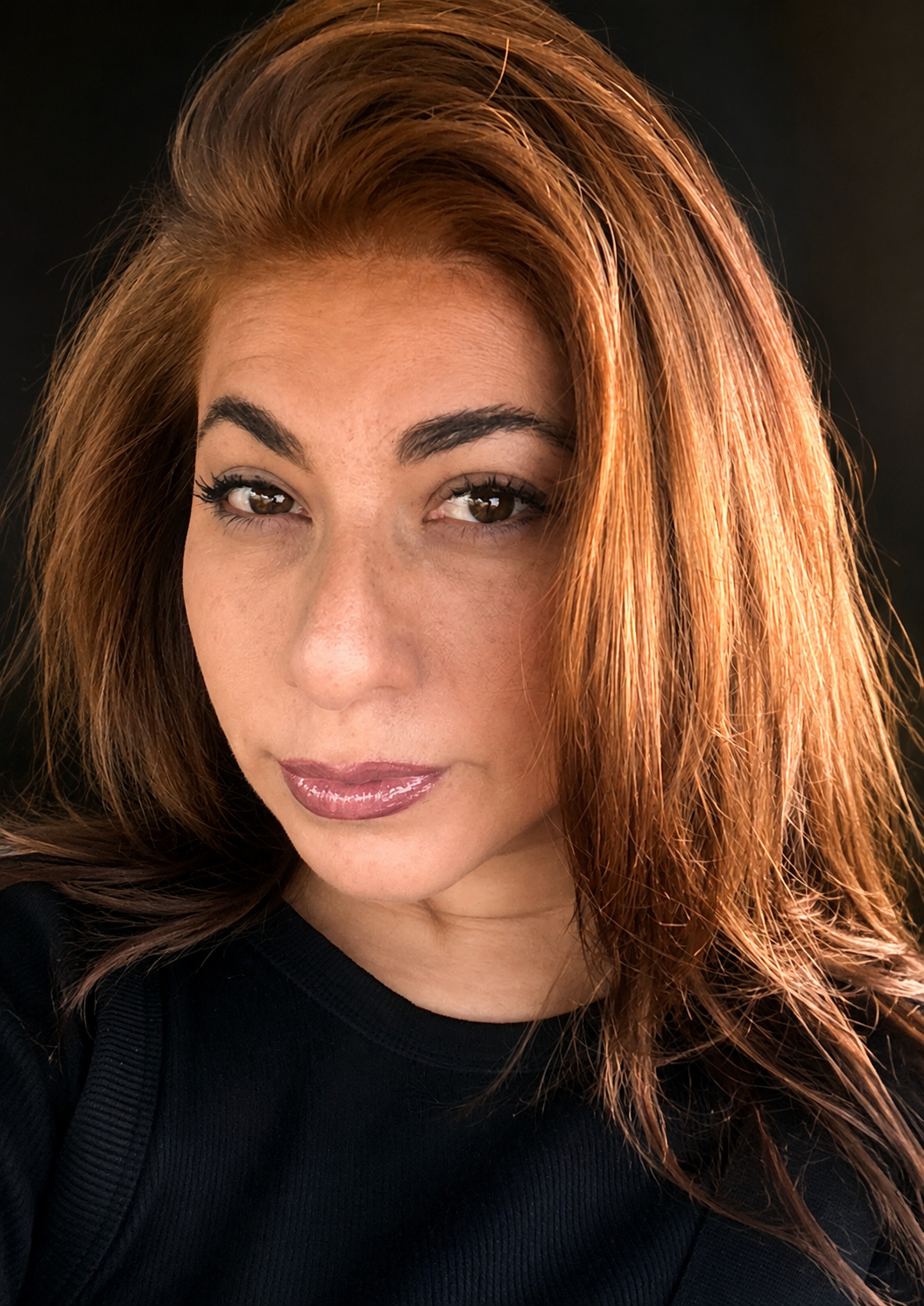 Close-up of a woman with red hair, light makeup, and a black top, looking at the camera against a dark background.