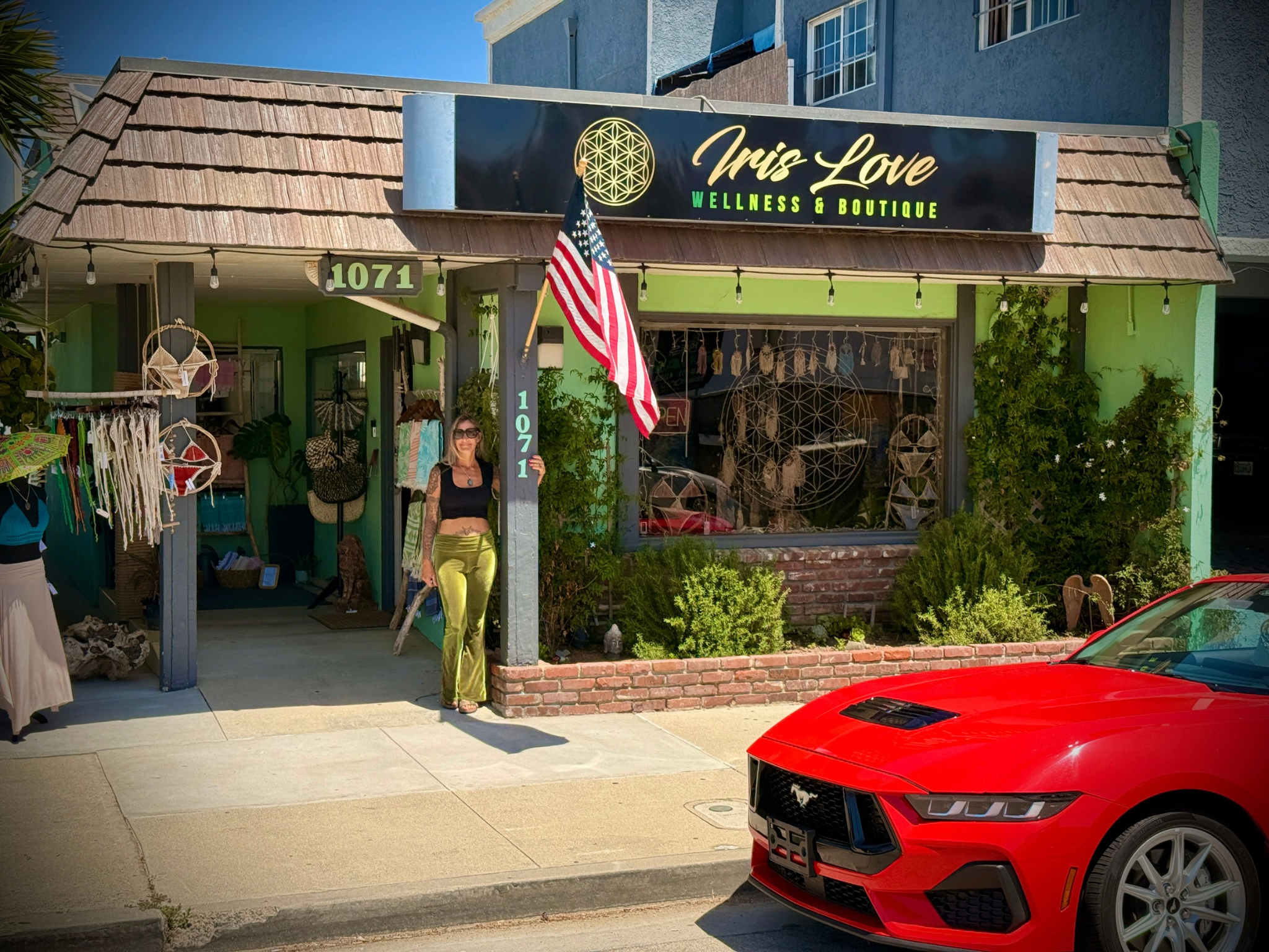 Storefront of Iris Love Wellness & Boutique with a woman holding an American flag, a red sports car parked in front, decorated with hanging lights, plants, and a display of home decor and clothing items.