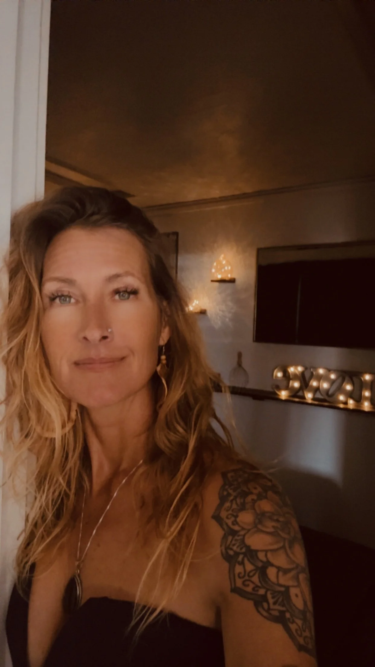 A woman with long, wavy blonde hair, wearing a black top and a necklace, taking a selfie in a dimly lit room with warm lighting and decorative items including light-up letters, a shelf, and wall art.