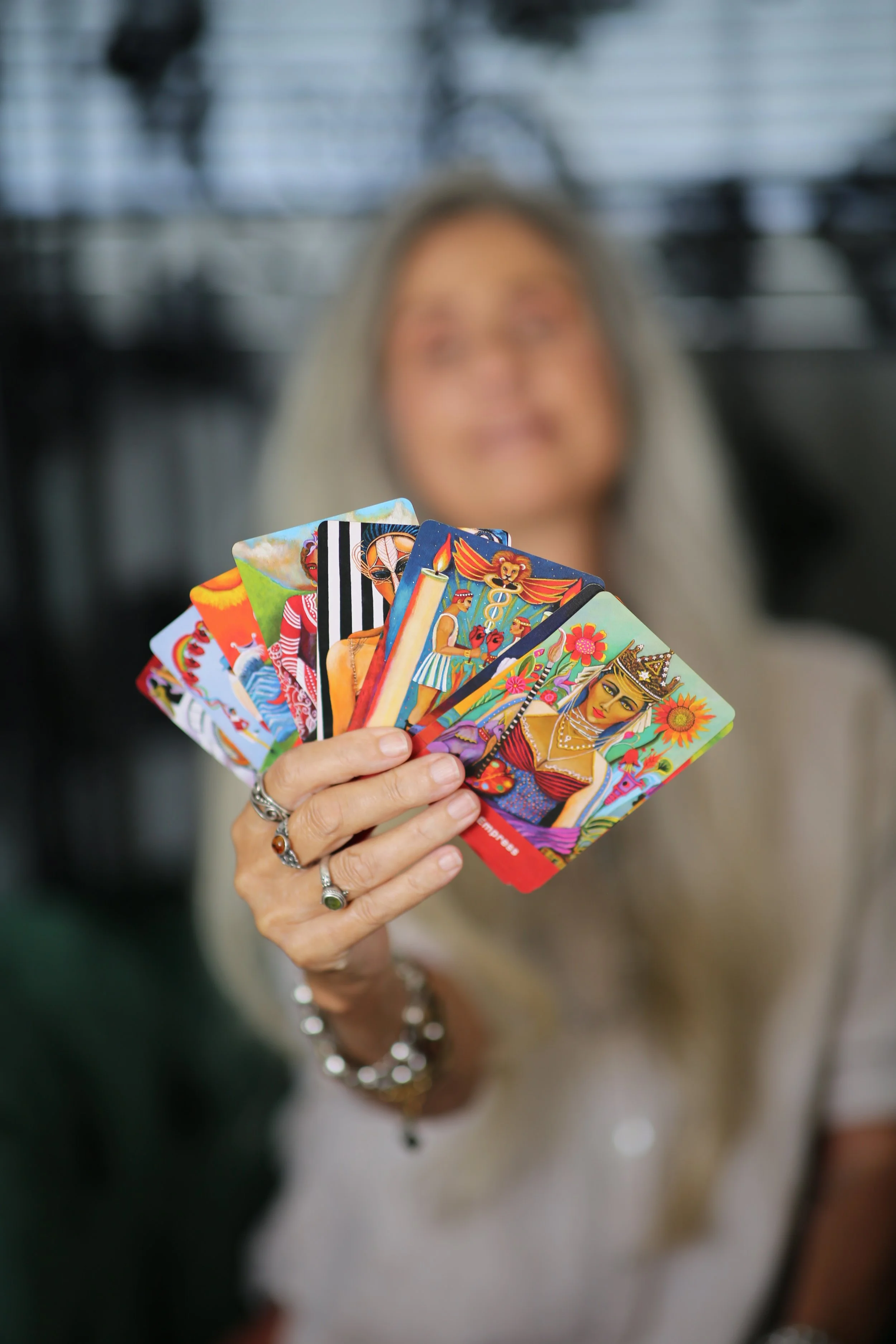 Anatasia, a  woman with long blonde hair wearing a colorful blue and yellow dress standing outside a wellness and bookshop store. The store has a sign promoting tarot readings today from 1 to 5, with decorative plants and a brick ledge in front.