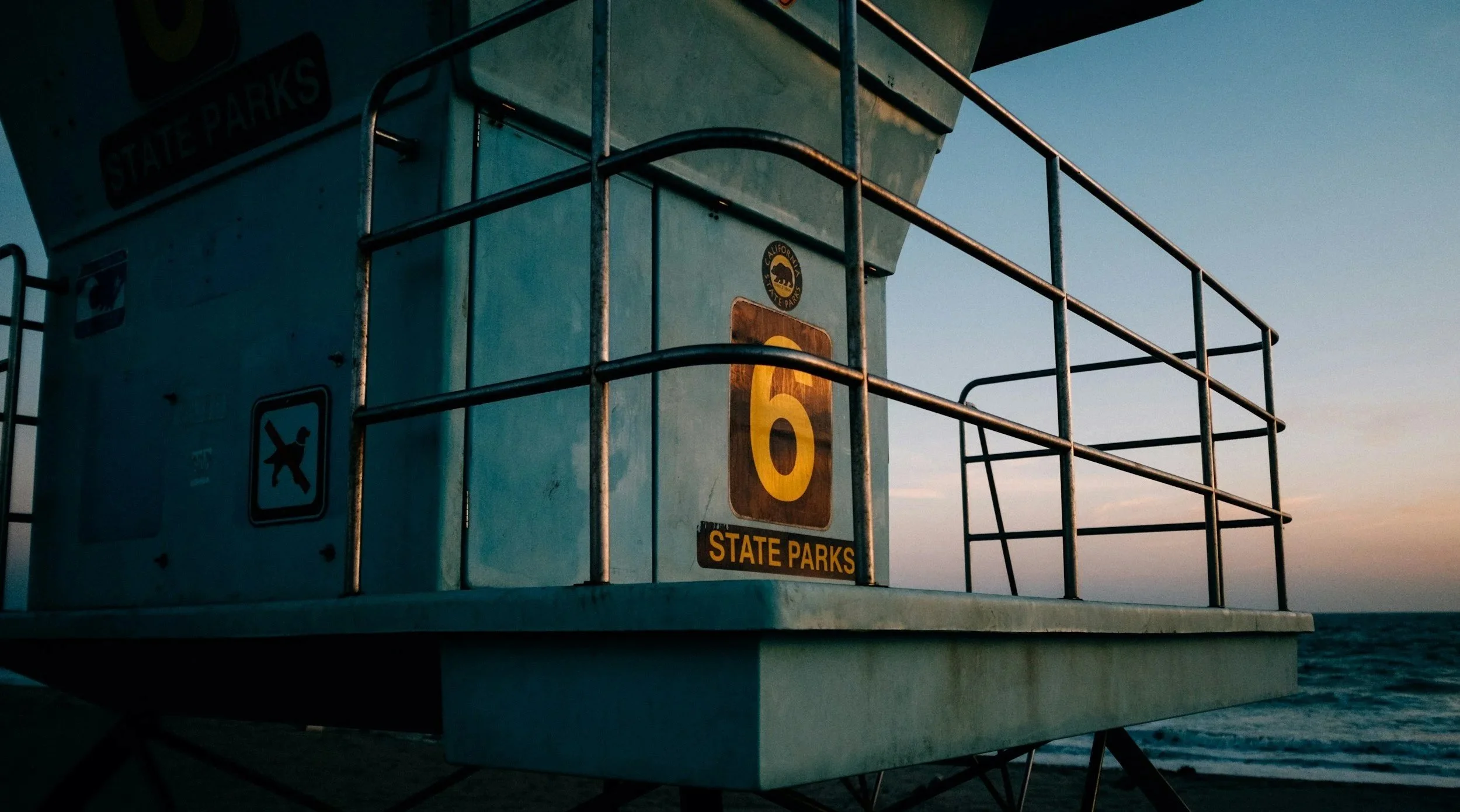 Lifeguard tower near the ocean at sunset with signage indicating state parks, a yellow number 6, and a black dog symbol.