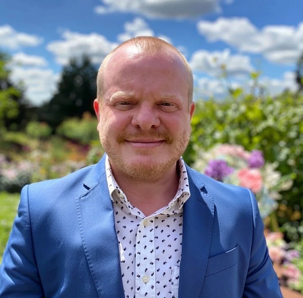 A man with a light beard and mustache wearing a light blue suit jacket and a white button-up shirt with small black patterns, standing outdoors with a background of a cloudy sky and blooming flowers.