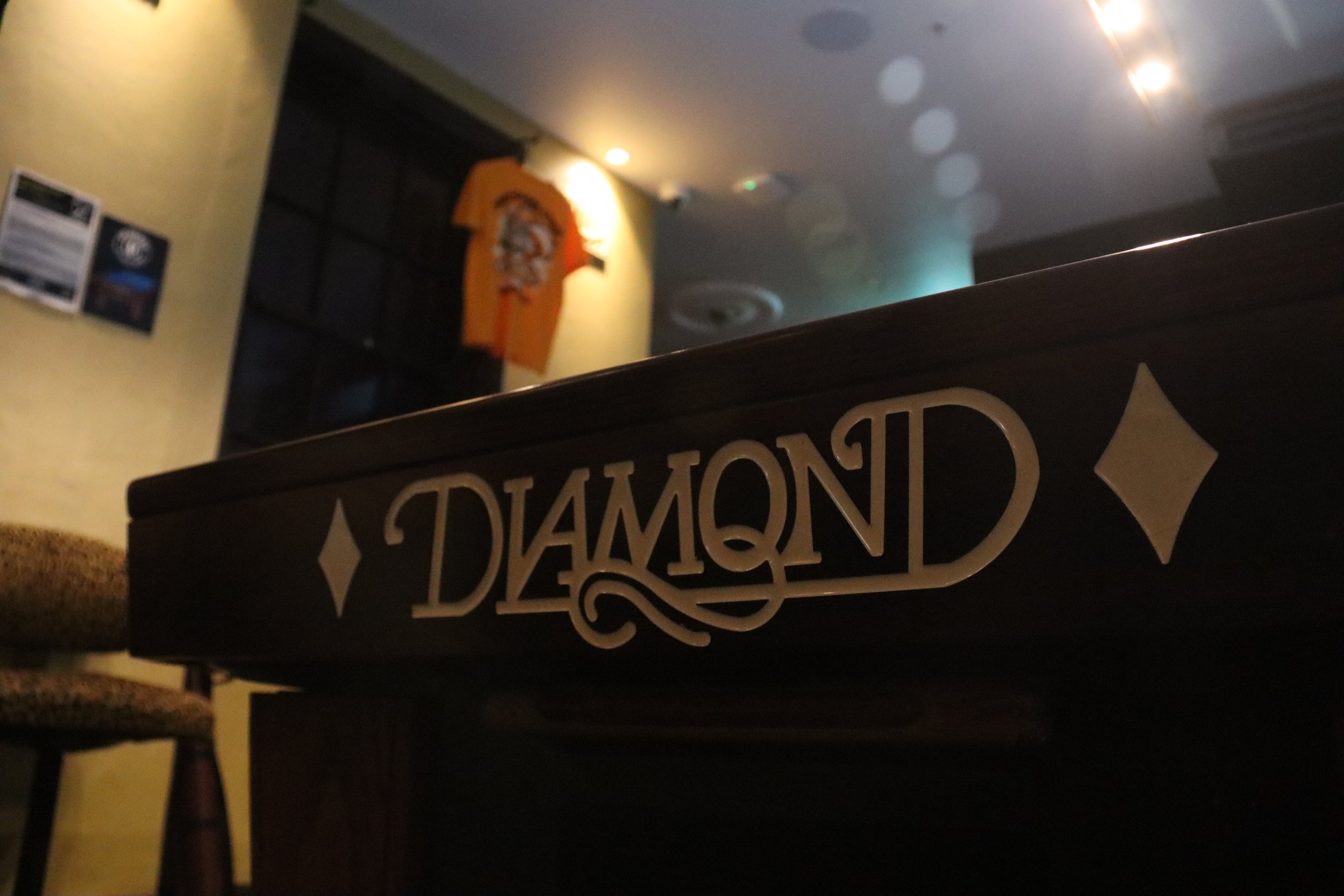 Close-up of a dark wooden billboard with the word 'DIAMOND' in stylized lettering and diamond symbols, dimly lit interior background with a wall, a window, and a hanging orange t-shirt.