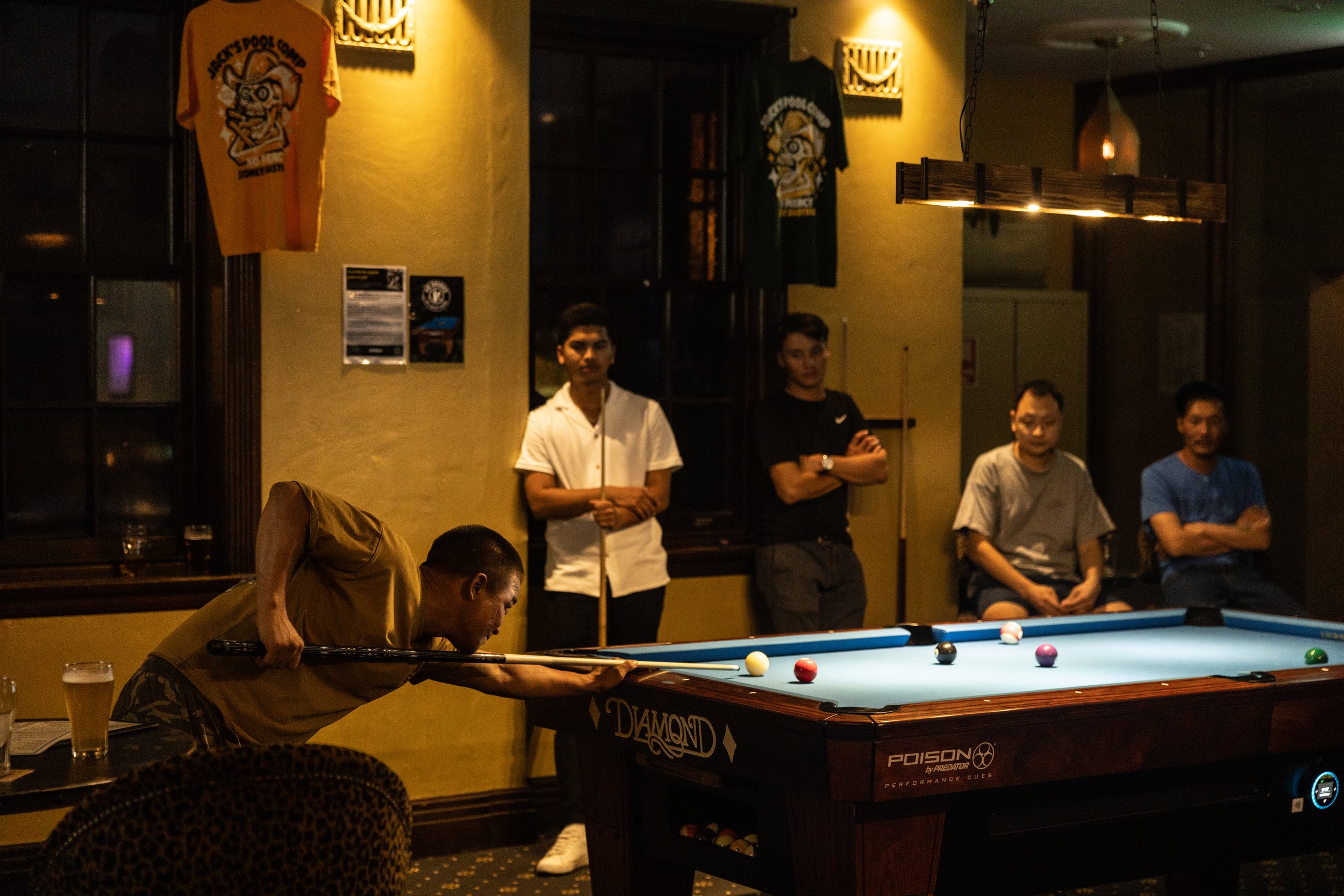 A man leaning over a pool table, aiming a shot with a cue stick. Several men are sitting or standing nearby watching, in a dimly lit room. A drink is on a nearby table. The wall displays pool-related items and logos.