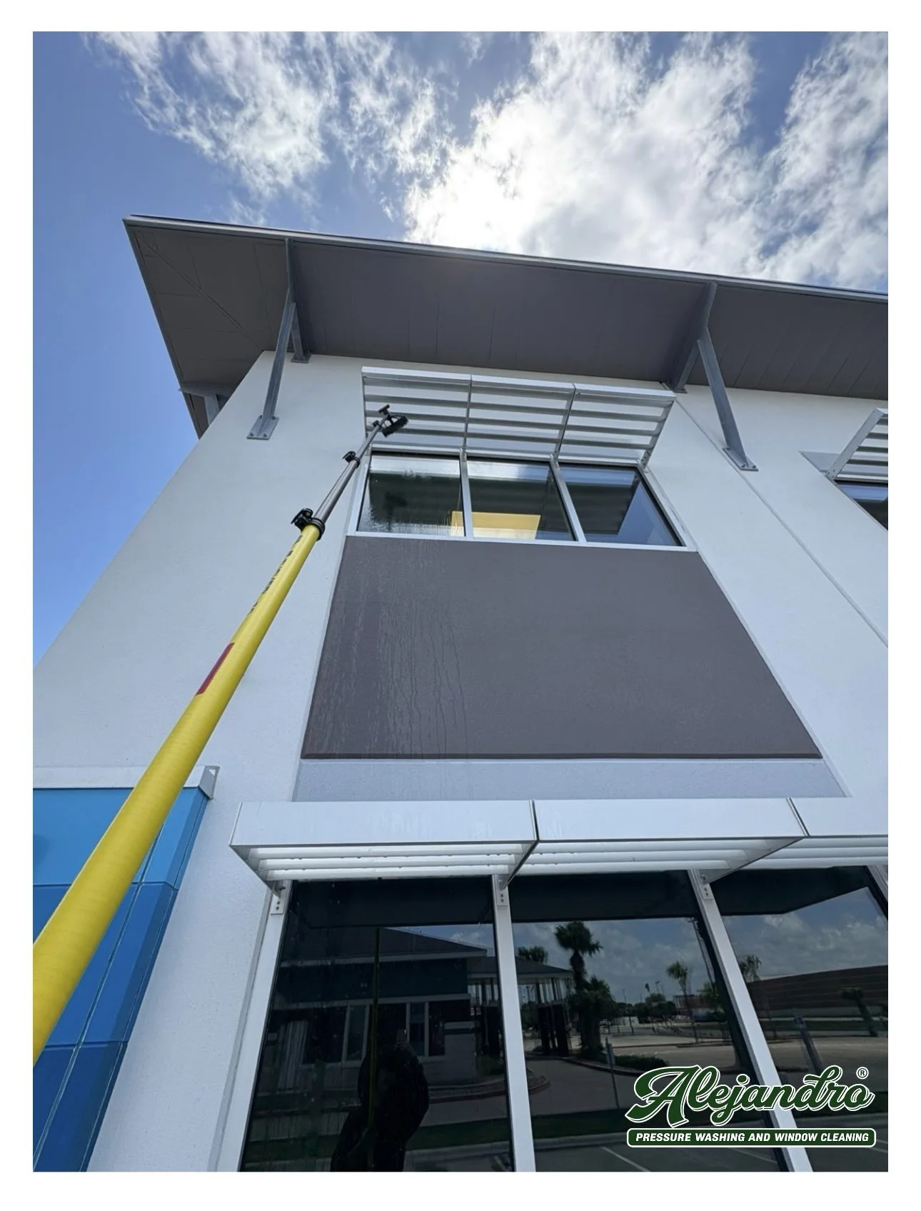 Low-angle outdoor view of a modern building with large glass windows and a dark gray exterior, under a blue sky with clouds, with a yellow pole and a window cleaning platform.