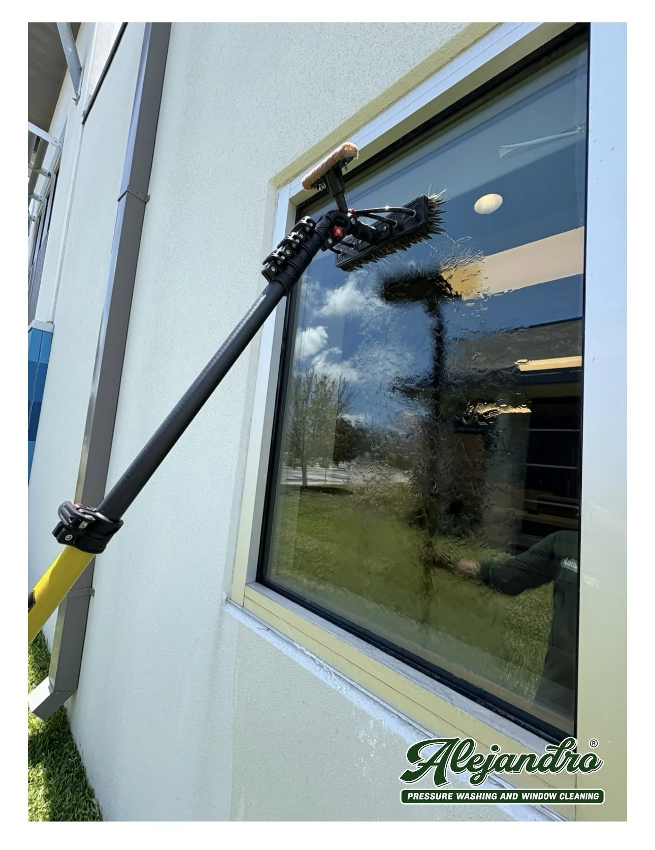 A pressure washing tool cleaning a large window on a building exterior with a white wall, trees, and sky reflected in the glass.