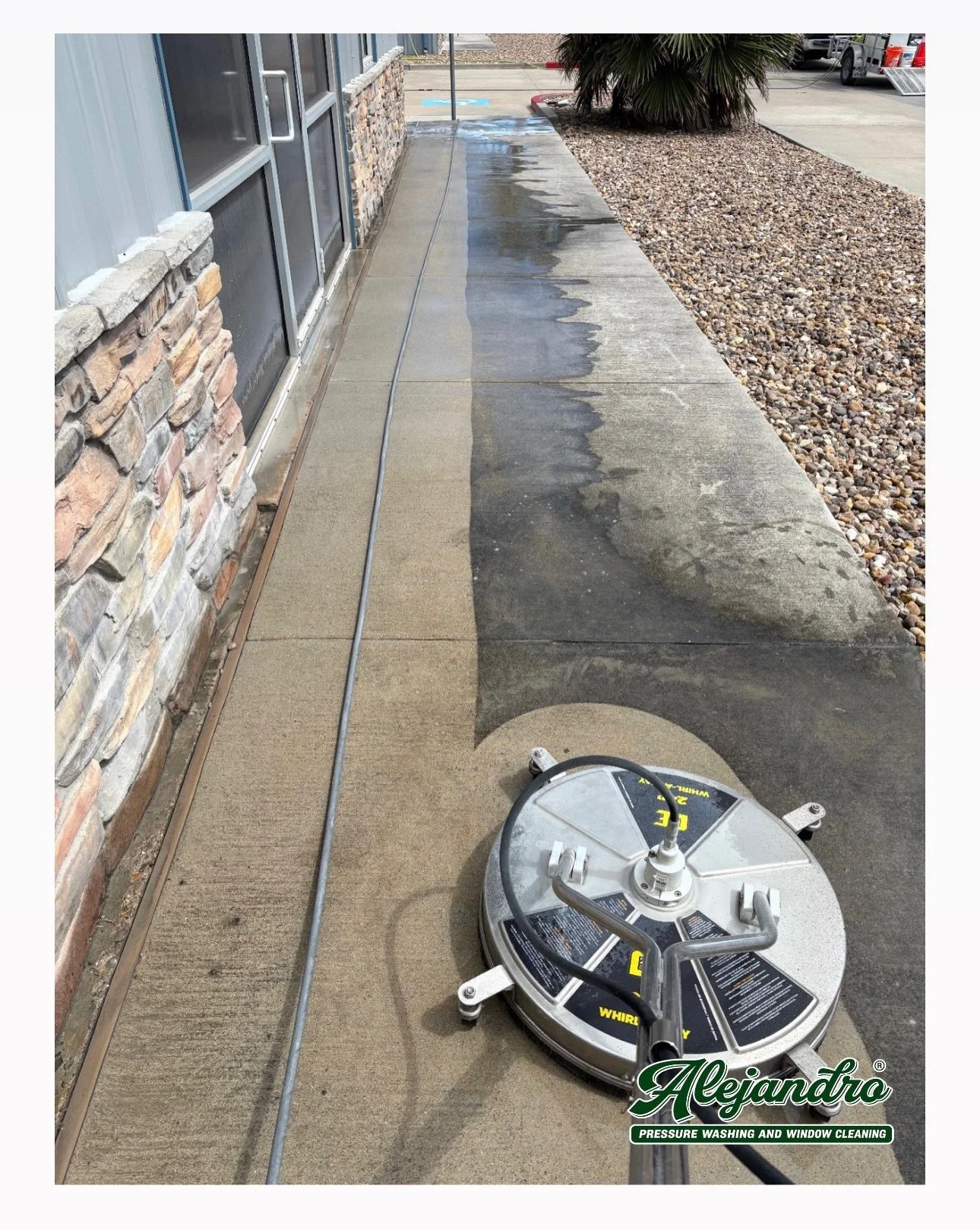 Pressure washing machine cleaning the concrete sidewalk outside a building with stone and brick exterior. Water is visible on the sidewalk, and a hose connects to the machine. A palm plant and parked vehicles are in the background.