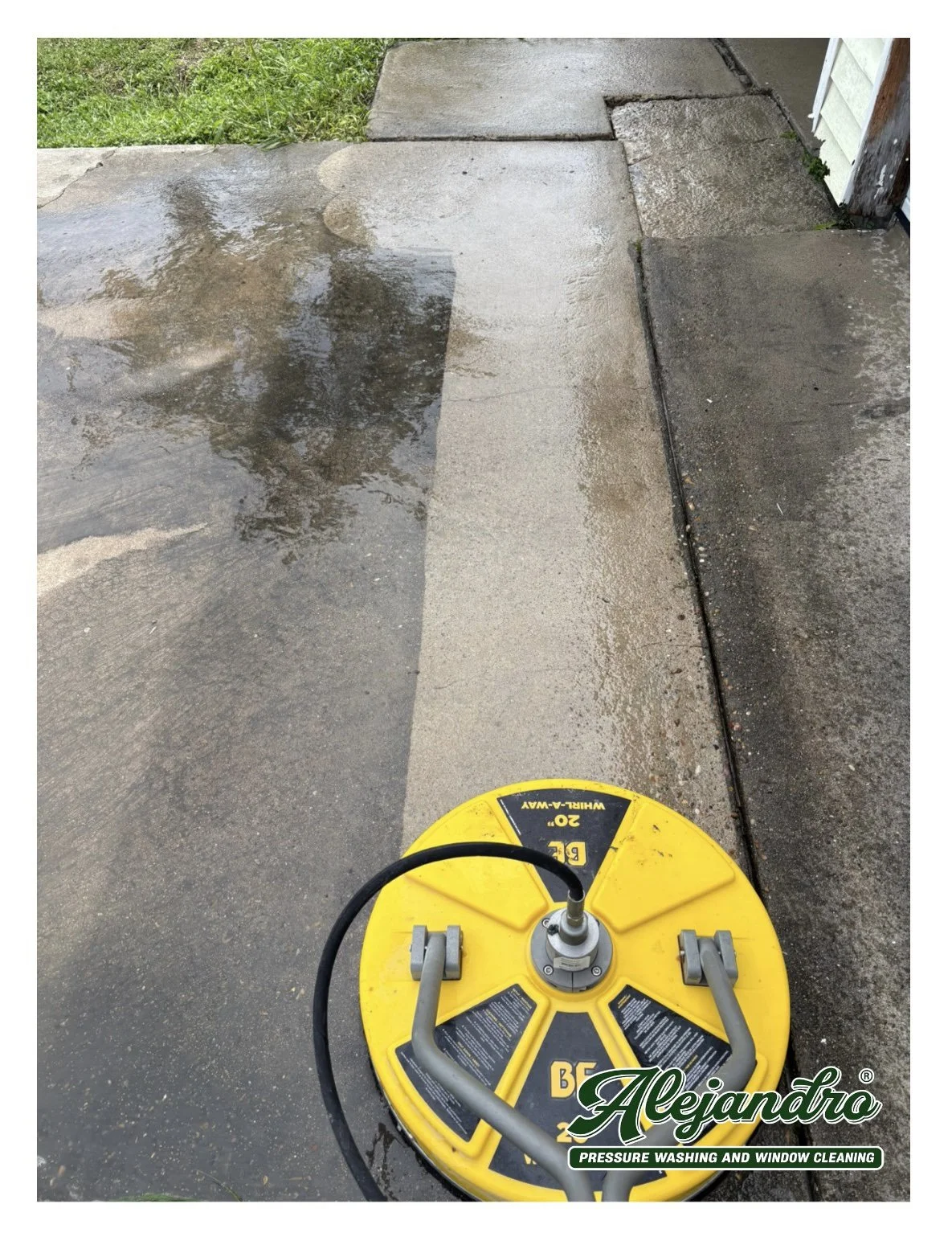 Wet concrete sidewalk with a pressure washing machine in the foreground and a grassy lawn on the left.