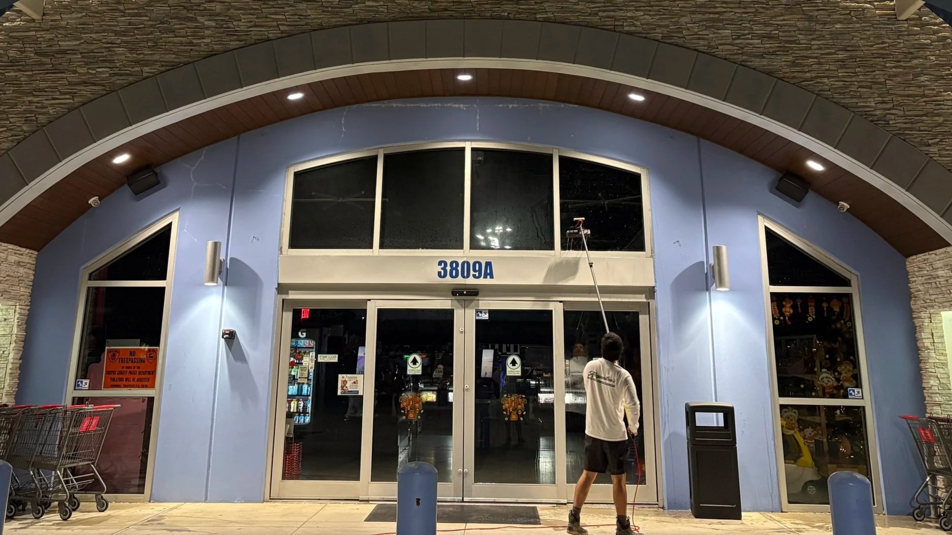 Person cleaning the exterior glass doors of a store at night using a cleaning tool with an extension pole. The store has blue walls, large glass windows, and a stone archway overhead, with shopping carts to the left.