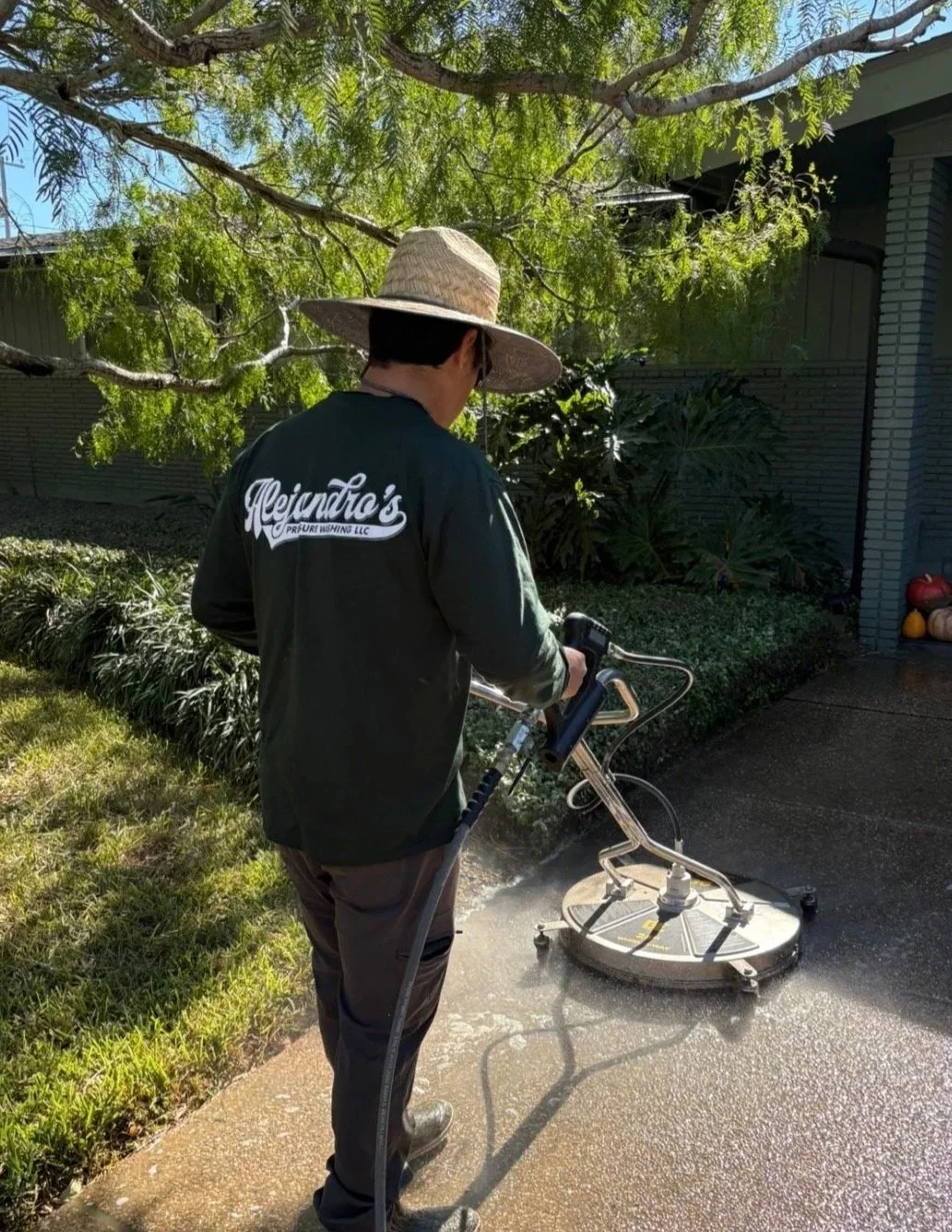 A person in a dark jacket and straw hat using a pressure washer to clean a concrete sidewalk during the daytime. There are trees and a house in the background.