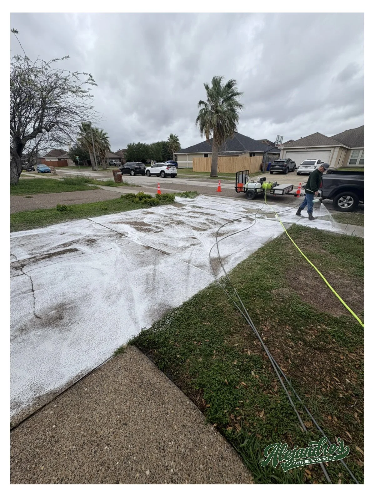 A person pressure washing a driveway outside a suburban house on a cloudy day. There are several parked cars and palm trees in the background.