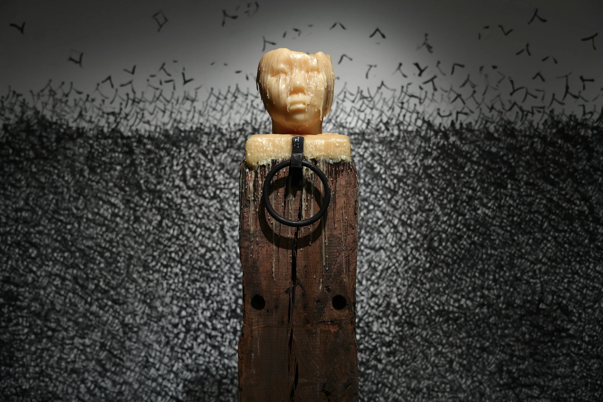 Referencing ancient Chinese Hitching post. A human head sculpture made of wax or similar material on a wooden pedestal, with a background of black staming character on wall and floor and a metallic ring hanging in front of the sculpture.