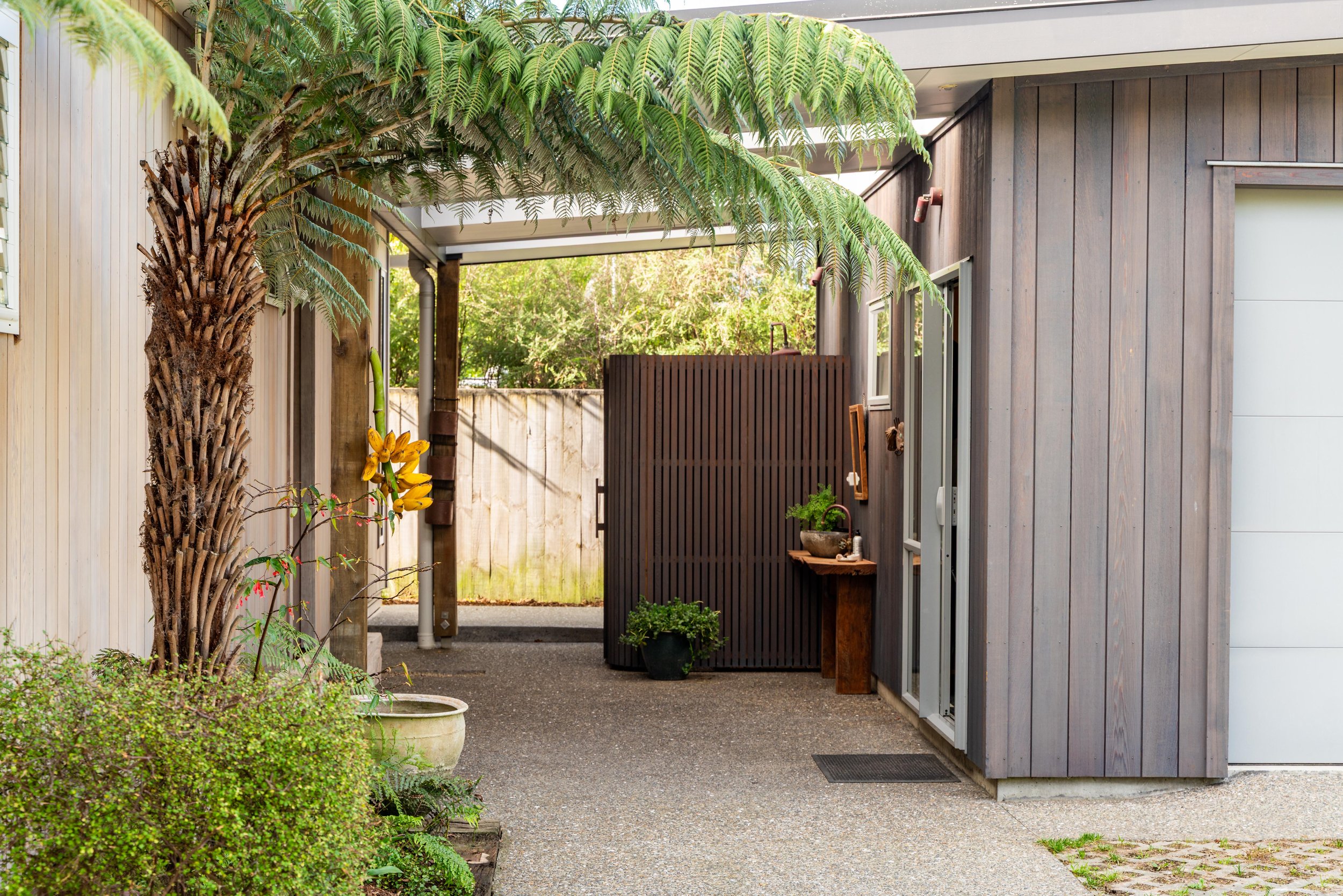 A side view of a modern house with gray wooden siding, featuring a small outdoor area with a palm tree, potted plants, and a wooden privacy screen.