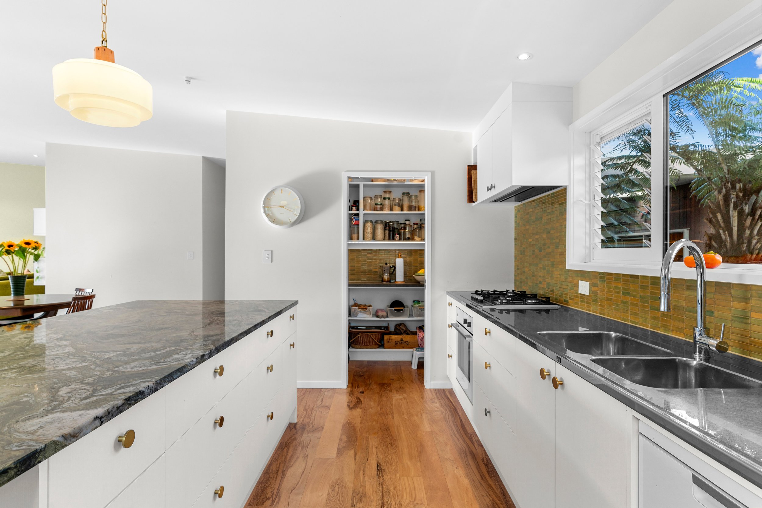 Modern kitchen with white cabinets, a granite countertop island, a double sink, a gas stove, and a window showing greenery outside. There is a pantry with jars of spices and dry goods and a wooden floor.