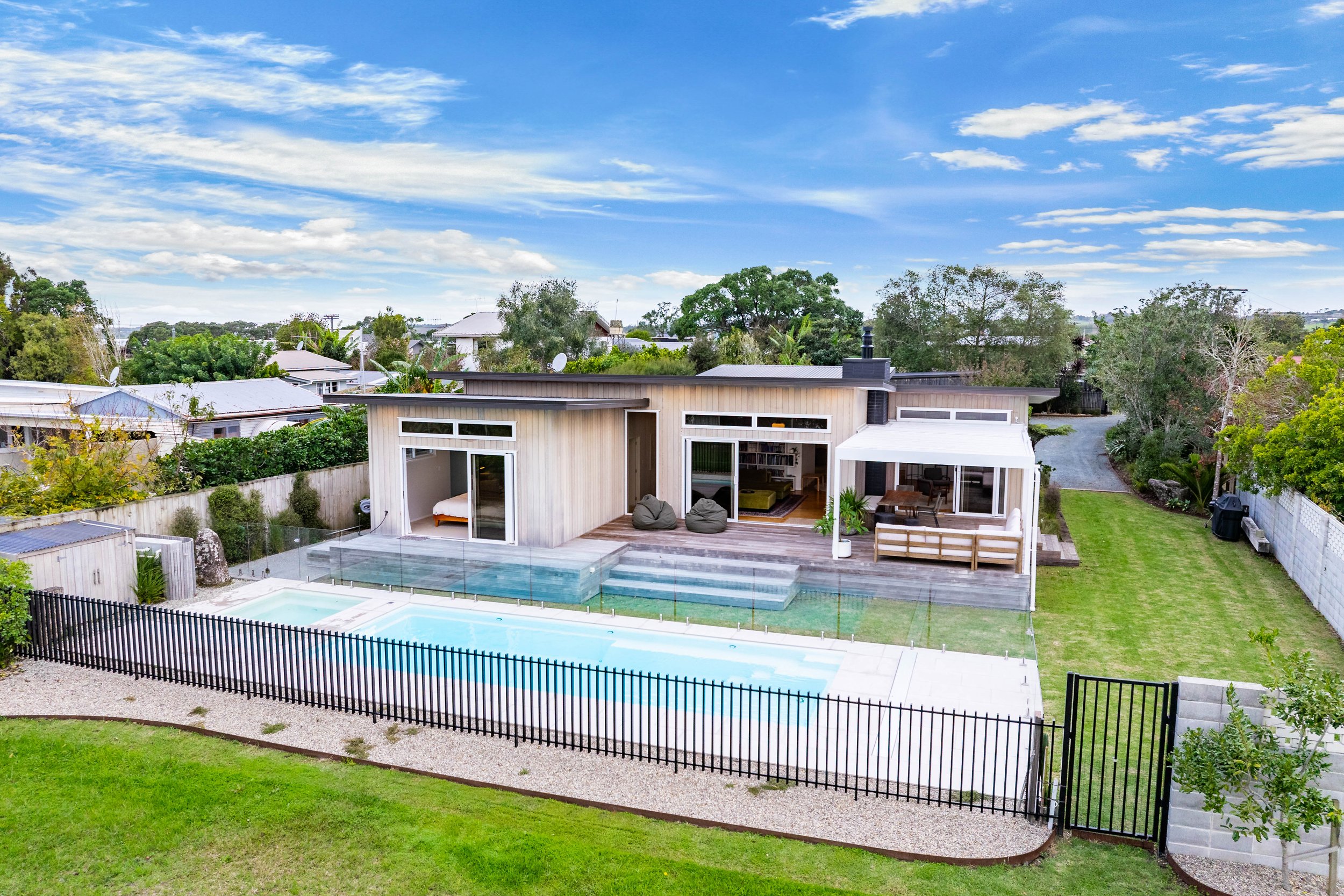 Modern house with wooden exterior, private swimming pool, and backyard surrounded by a black metal fence, with trees and neighboring houses in the background under a partly cloudy sky.