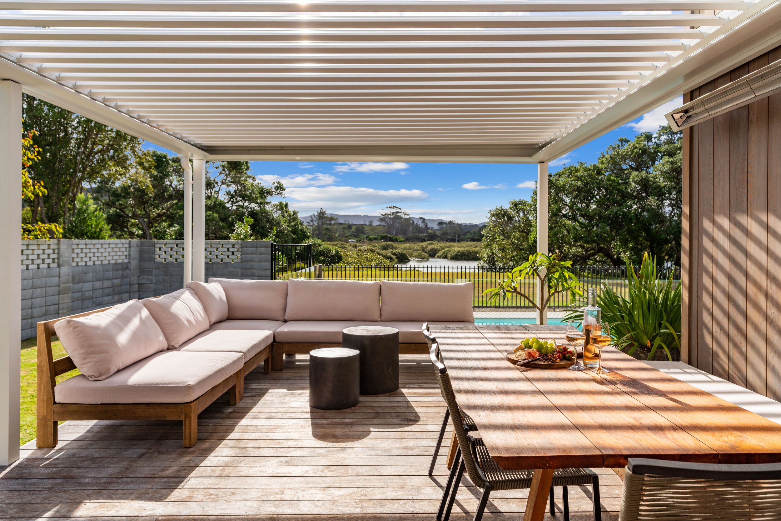 Outdoor patio with a wooden dining table, lounge seating, and a scenic view of trees, a pond, and a partly cloudy sky.