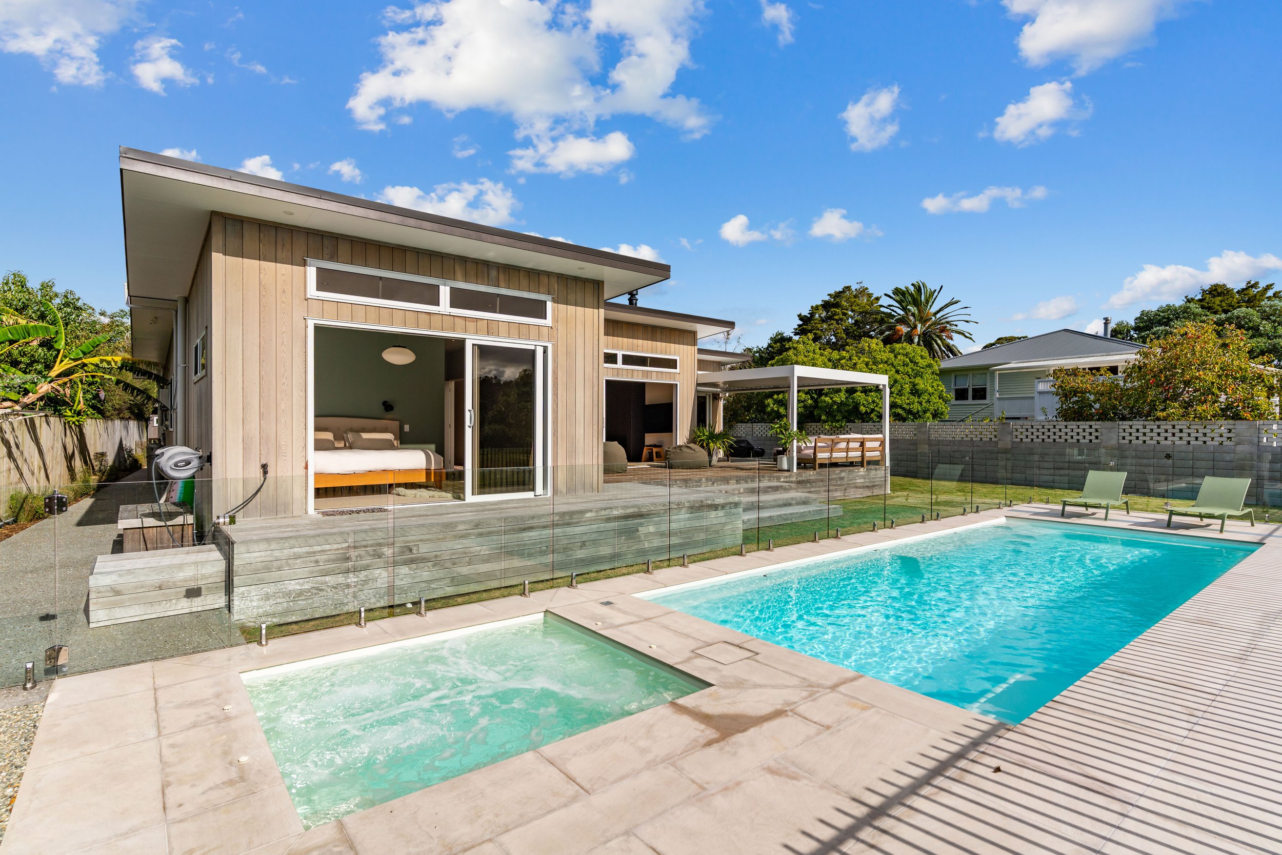 Modern house with large backyard, swimming pool, hot tub, outdoor lounge area, and glass fence under a blue sky with scattered clouds.