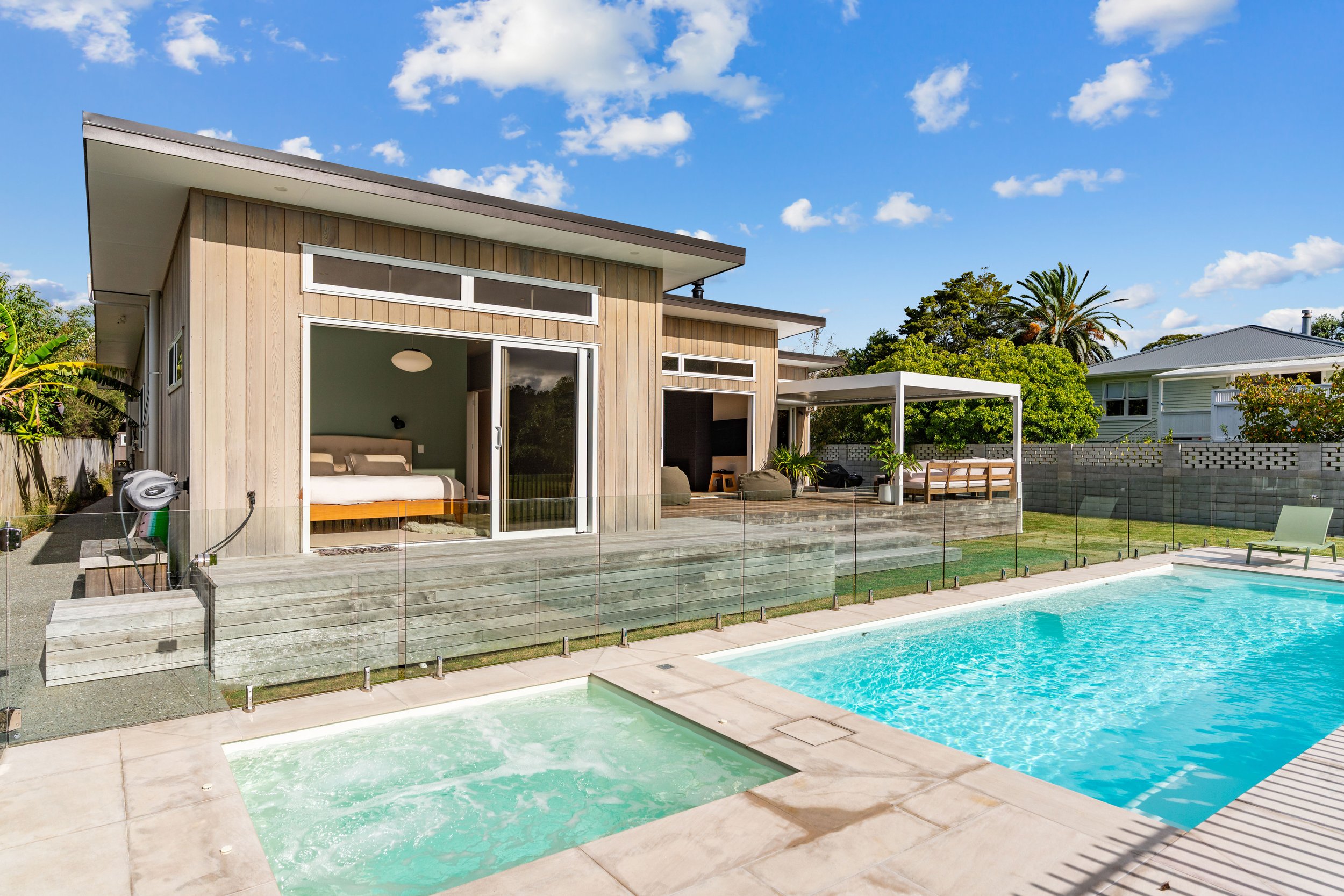 Modern house with wooden exterior, outdoor swimming pool, hot tub, patio furniture, and landscaped yard under a partly cloudy sky.