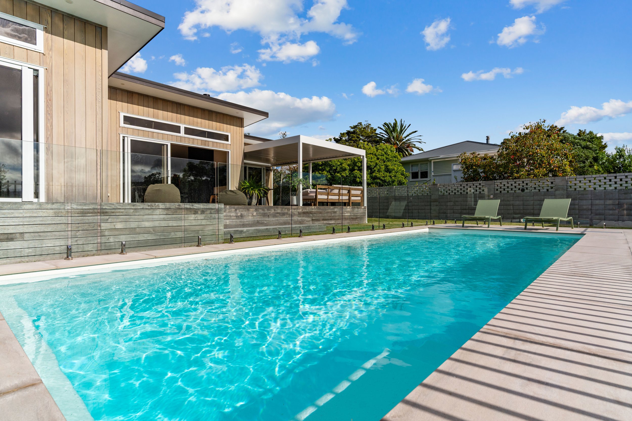 A backyard swimming pool with clear blue water, lounge chairs on the pool deck, and a modern house with large windows and a patio in the background, with trees and a partly cloudy sky.