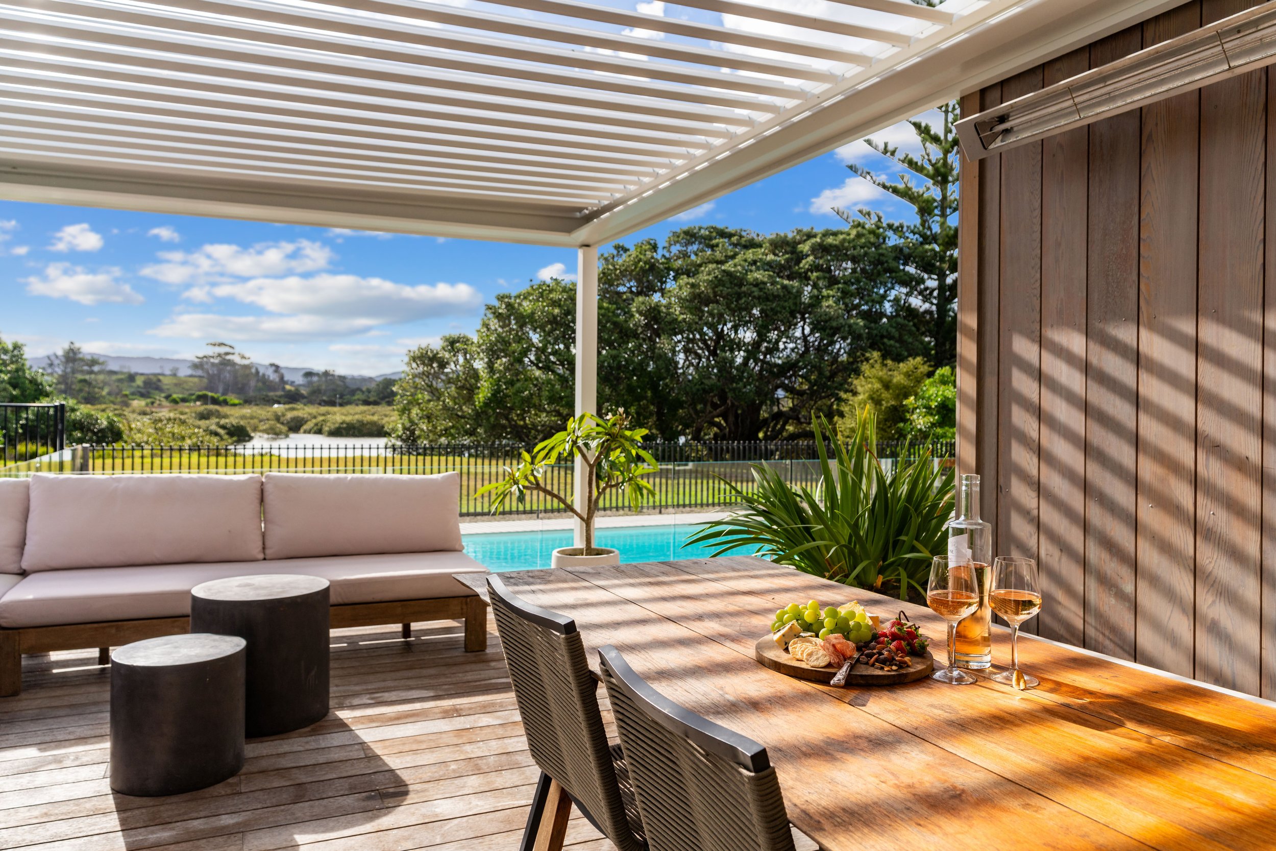 Outdoor patio with a wooden dining table, wine glasses, and a bottle, overlooking a swimming pool and lush green trees under a partly cloudy sky.
