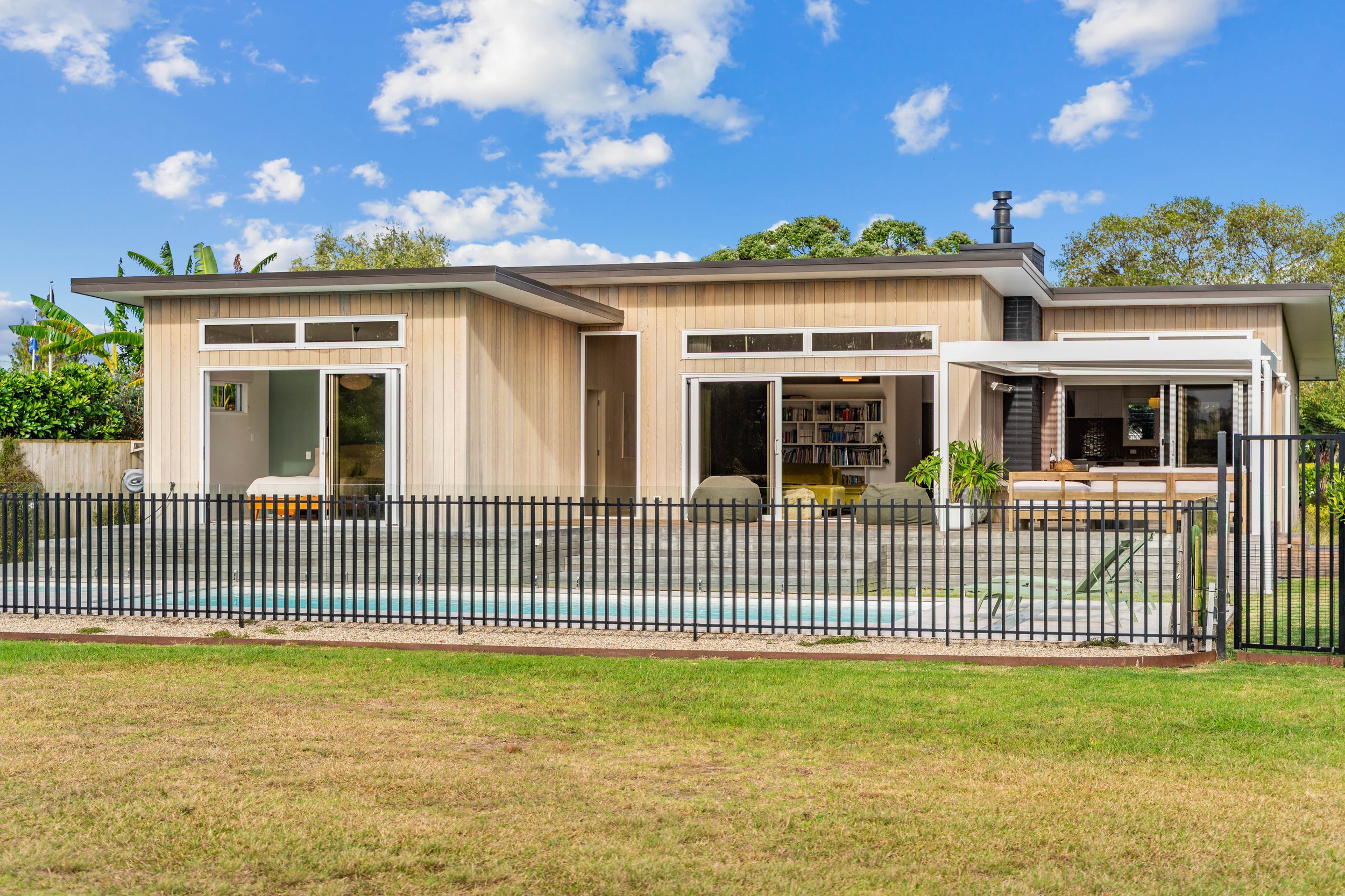 Modern house with a backyard, black safety fence, patio, and a swimming pool, under a blue sky with scattered clouds.