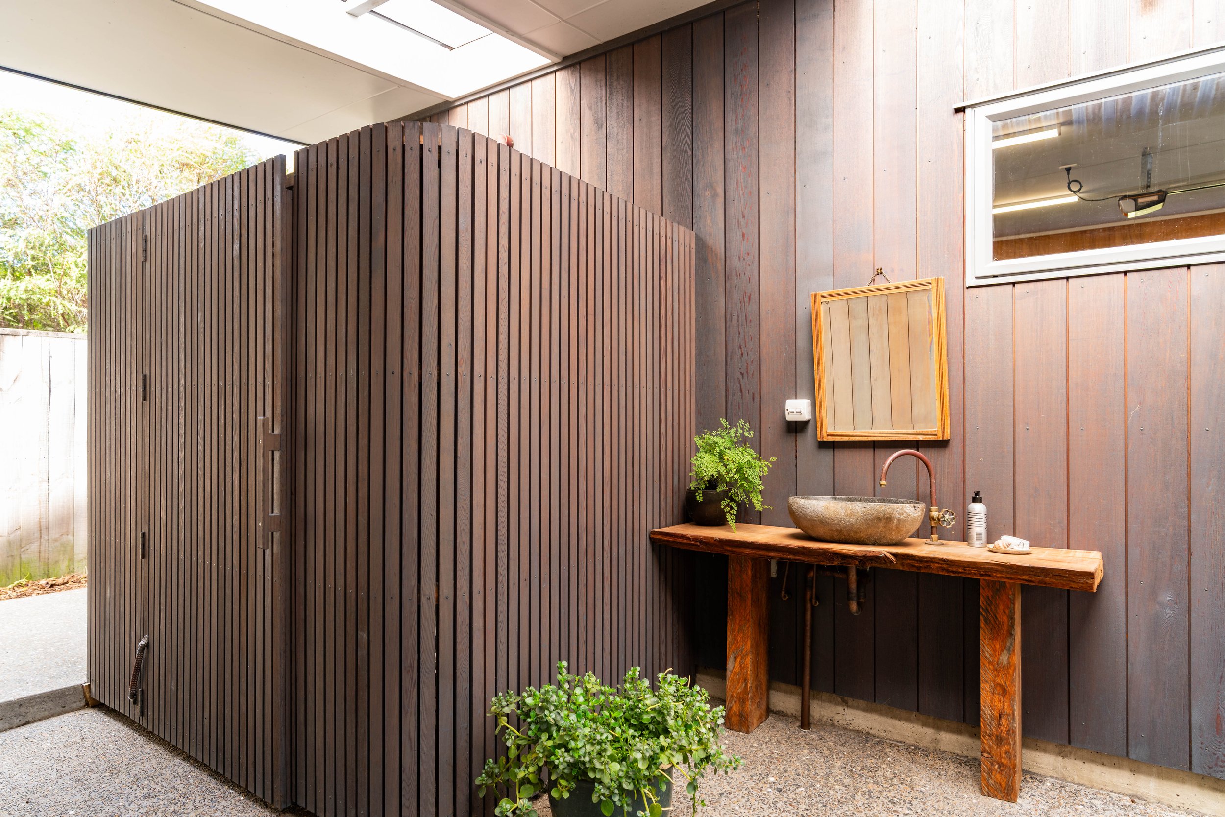 Outdoor bathroom with a large wooden shower enclosure, a rustic wooden vanity with a stone sink, a small mirror, potted plant, soap dispenser, and a window on a dark wooden wall.