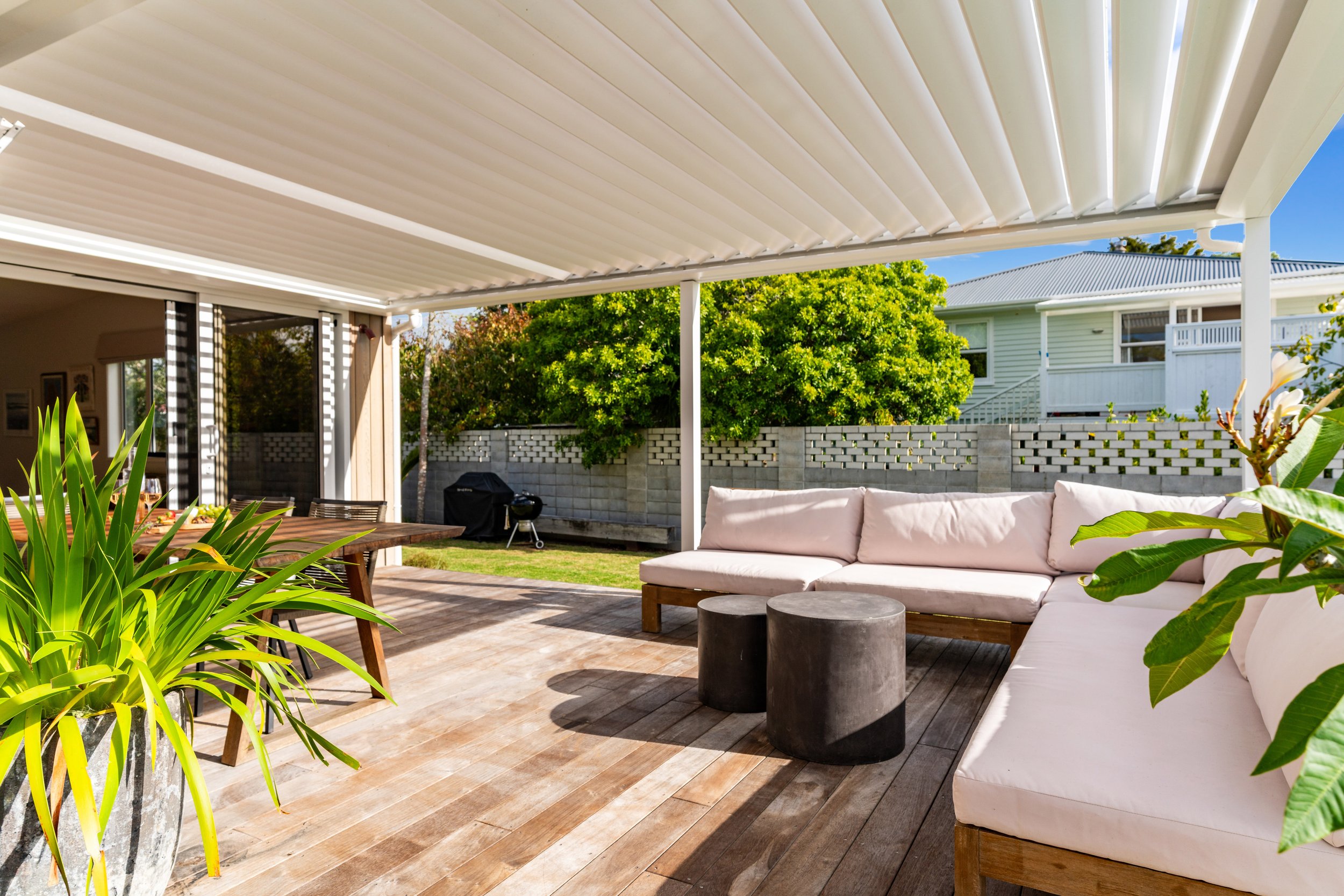 Outdoor patio area with a large L-shaped white cushioned sectional, black cylindrical side tables, wooden dining table with chairs, potted plants, a grill, and a backyard with a concrete block fence and greenery under a covered roof.