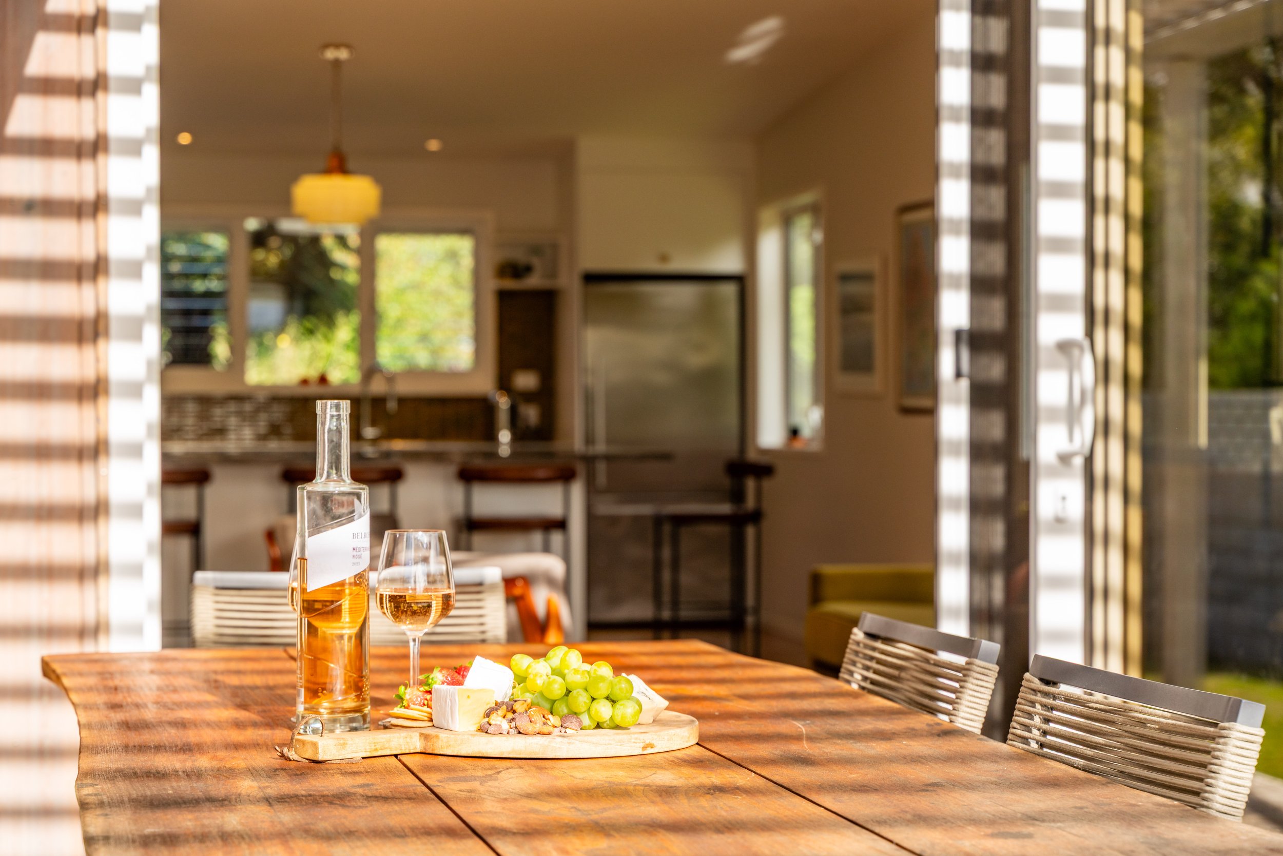 A wooden dining table with a bottle of rosé wine, a glass of rosé wine, and a cheese and fruit platter, set in a sunlit kitchen or dining area with open doors and windows.