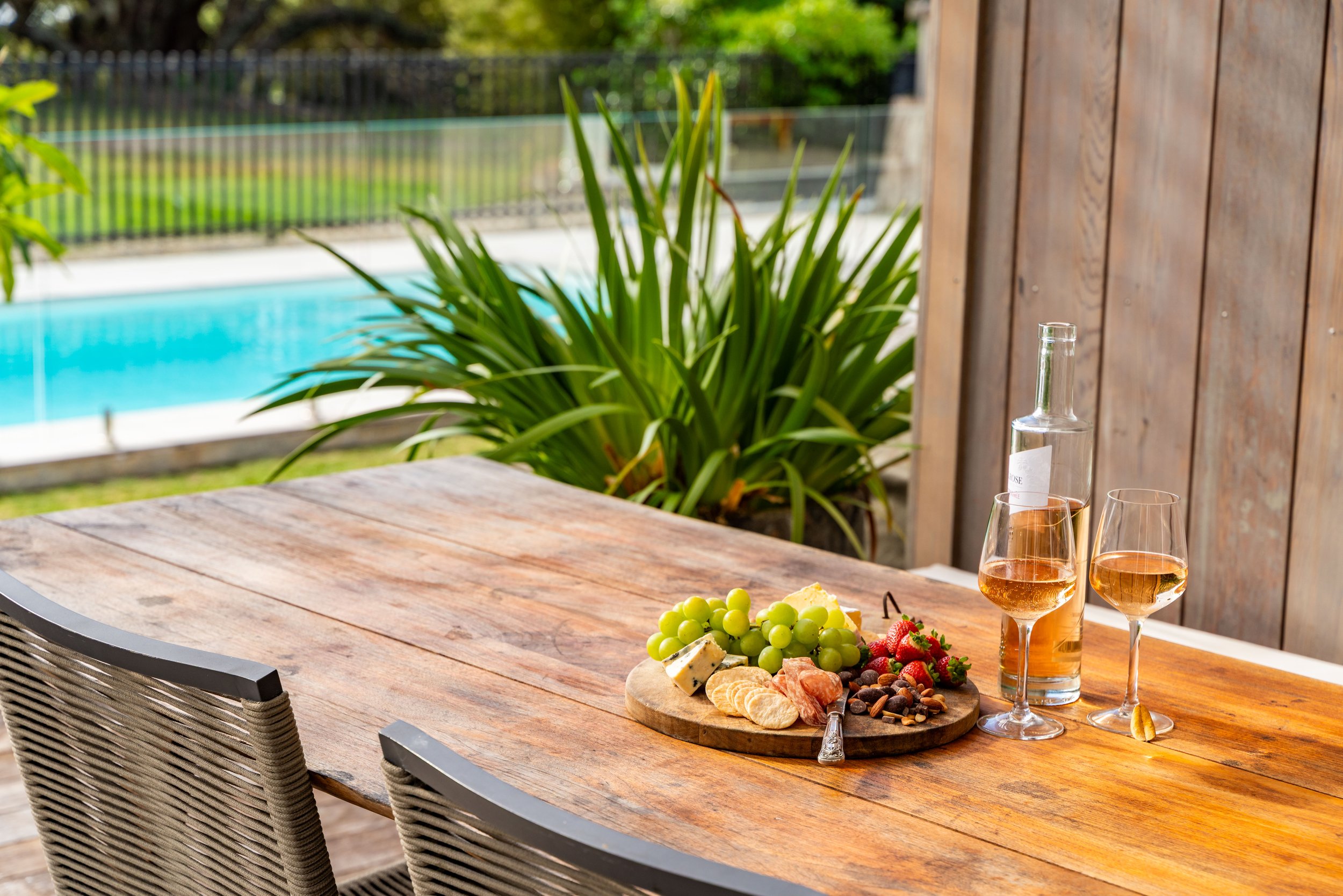 Outdoor wooden table with a cheese and fruit platter, two glasses of rosé wine, and a clear glass bottle, near a pool and green plants.