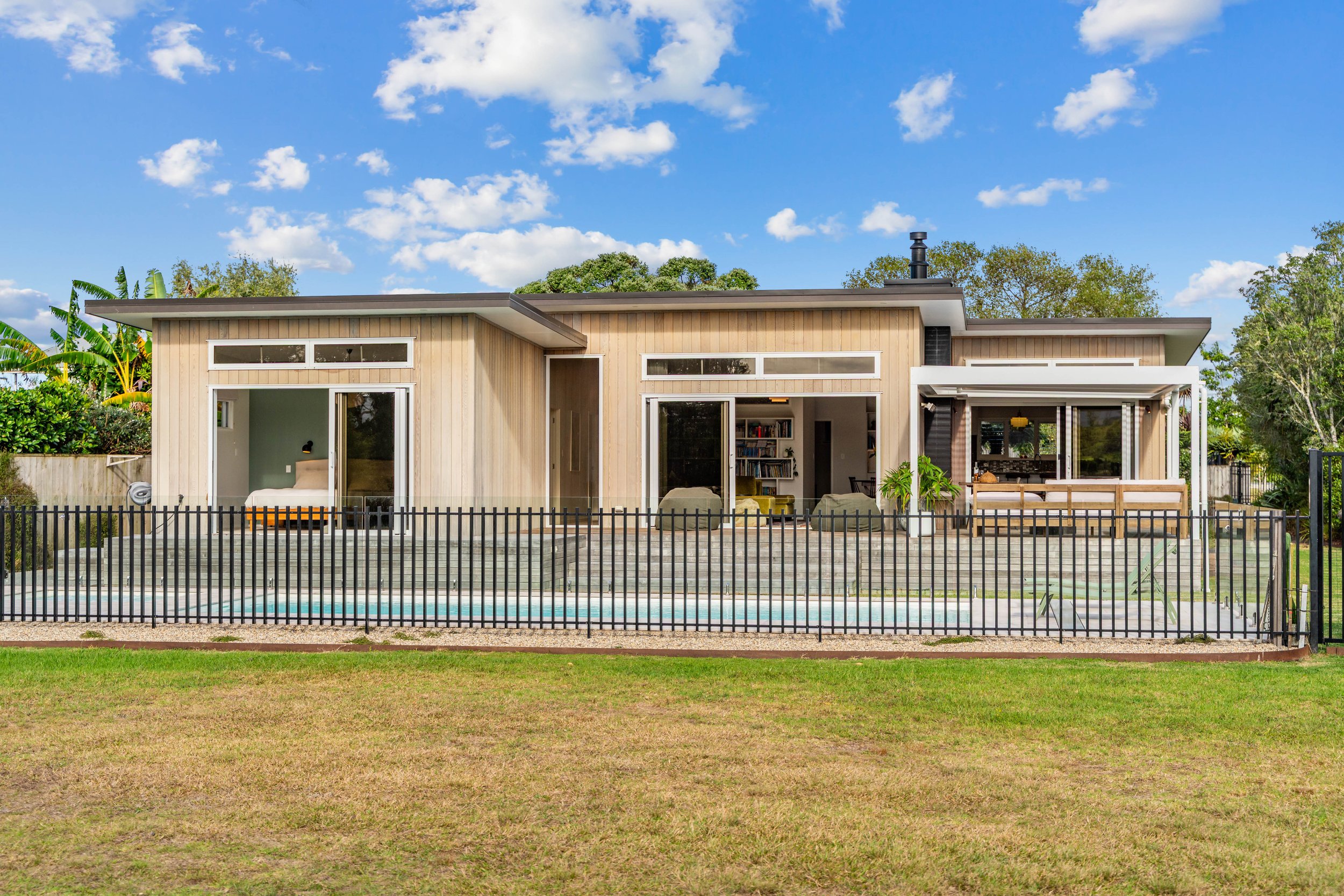 Modern house with wooden exterior, large sliding glass doors, small backyard pool, black metal fence, outdoor seating area, and lush greenery under a bright blue sky.