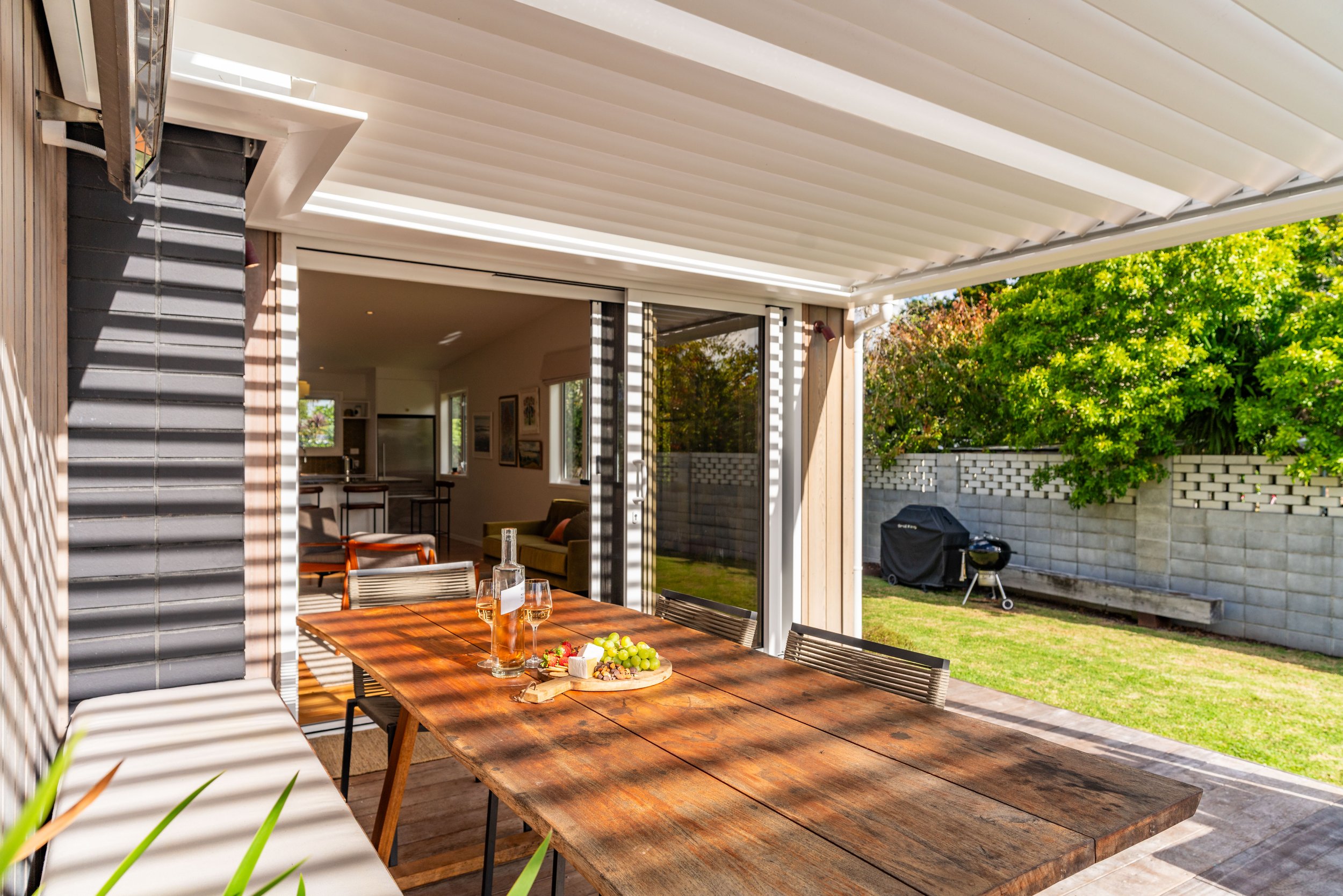 Outdoor patio with wooden dining table, chairs, and sunshade, connected to indoor living space with windows, yard with trees, grill, and wooden bench.