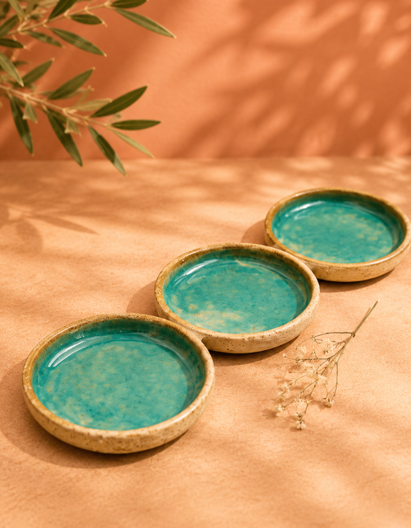 Three small ceramic bowls with blue-green glaze on a peach-colored surface, with a sprig of dried flowers and a leafy branch nearby.